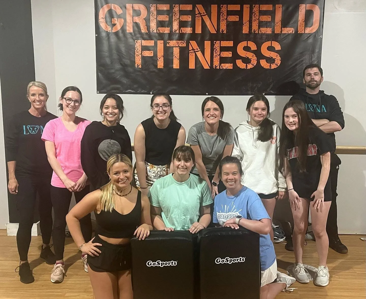 Group of women and men at Greenfield Fitness, standing and kneeling indoors with fitness equipment, group photo for a fitness class or team.