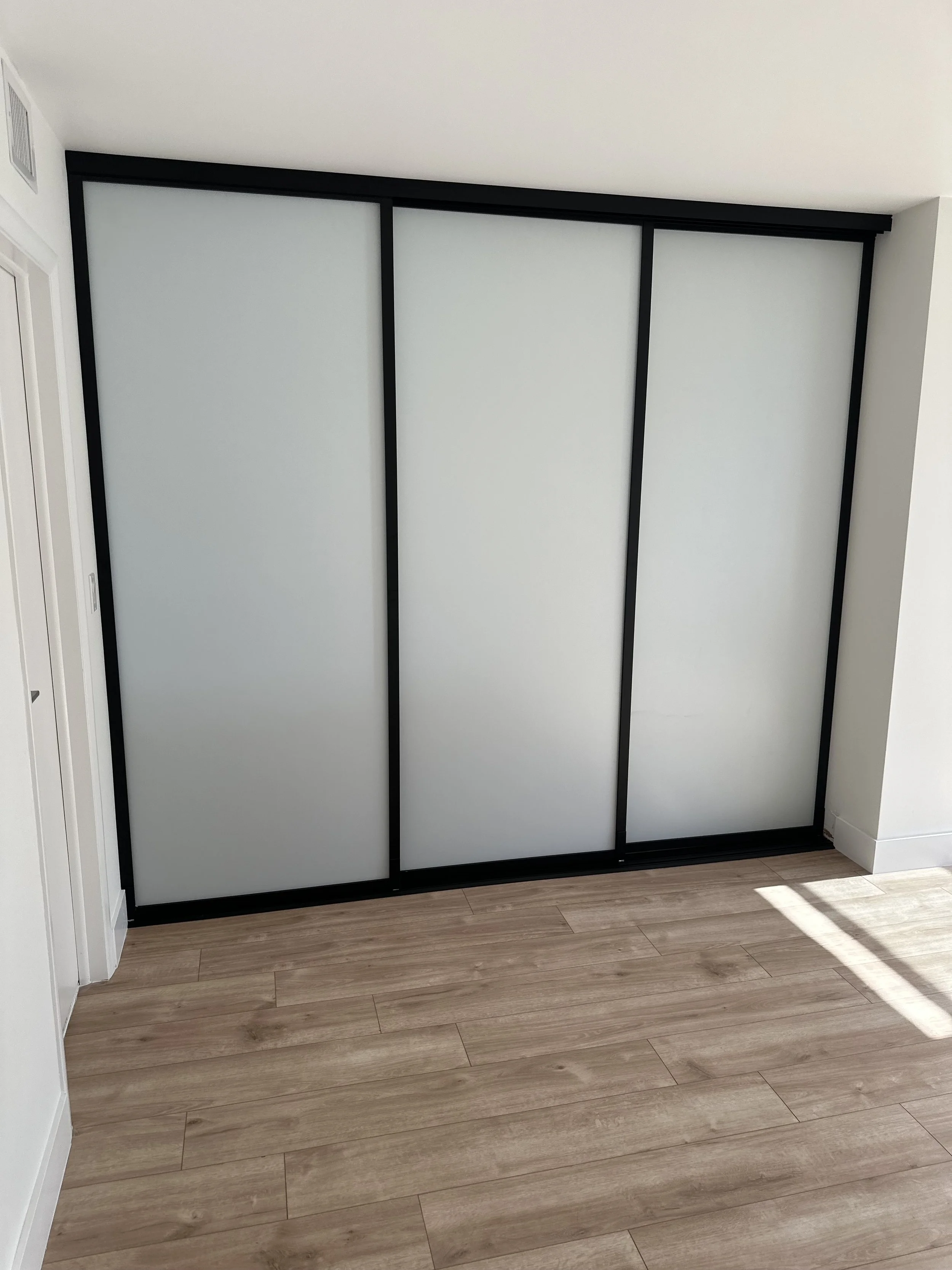 A closet with sliding frosted glass doors framed in black, set in a room with light wood flooring and white walls.