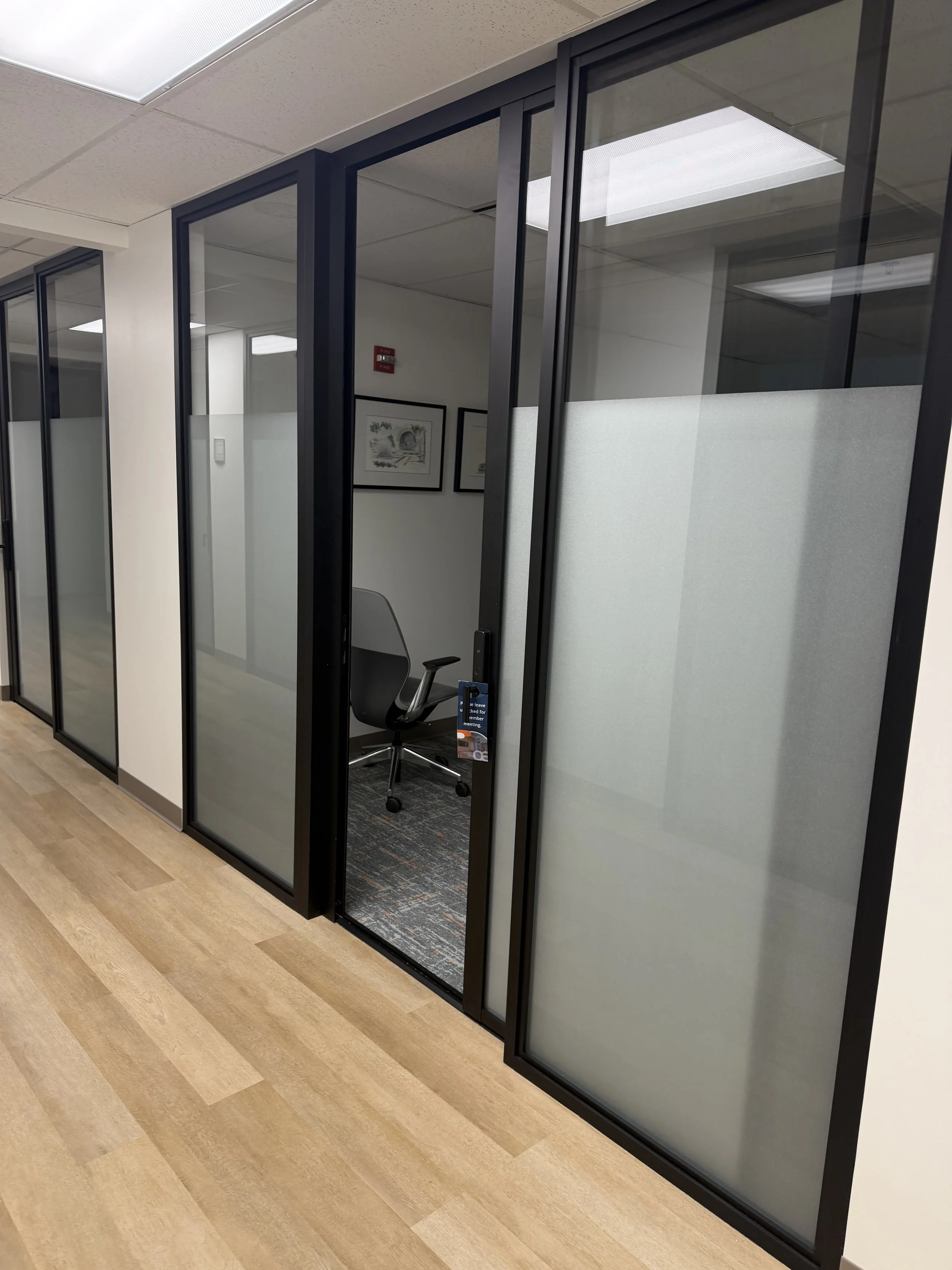Empty office cubicle with frosted glass walls, a black office chair, and wall art, in a modern office hallway with wood flooring.