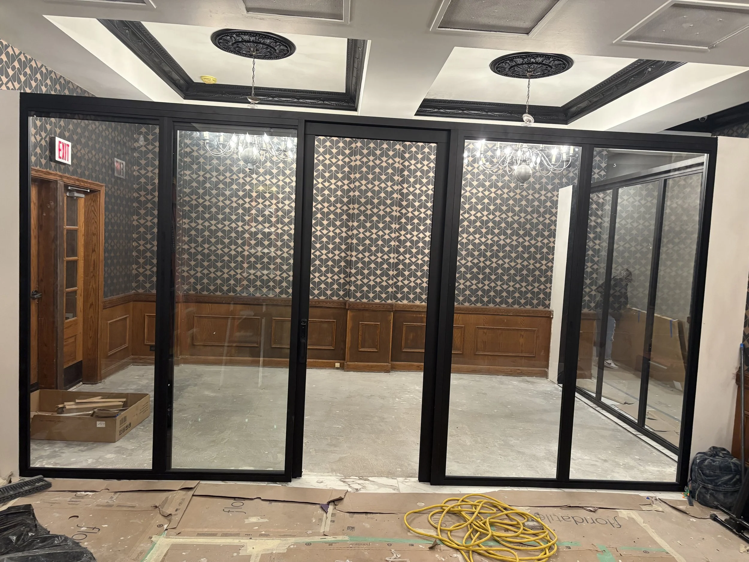 Interior space under construction with glass partitions, patterned wallpaper, and wood wainscoting, with construction materials and tools on the floor.