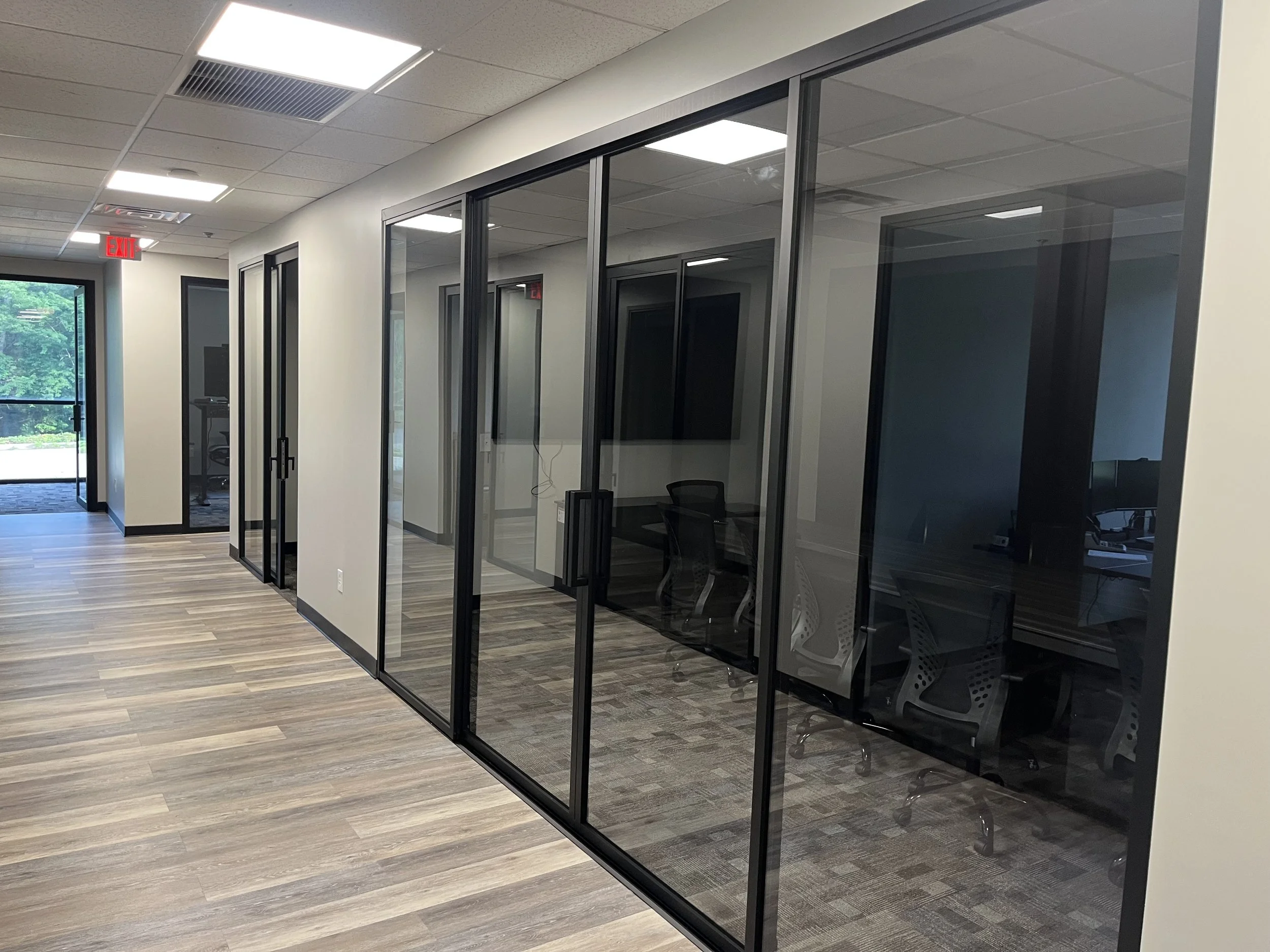 A modern office hallway with glass-walled conference rooms, wood flooring, and an exit door with an emergency exit sign.