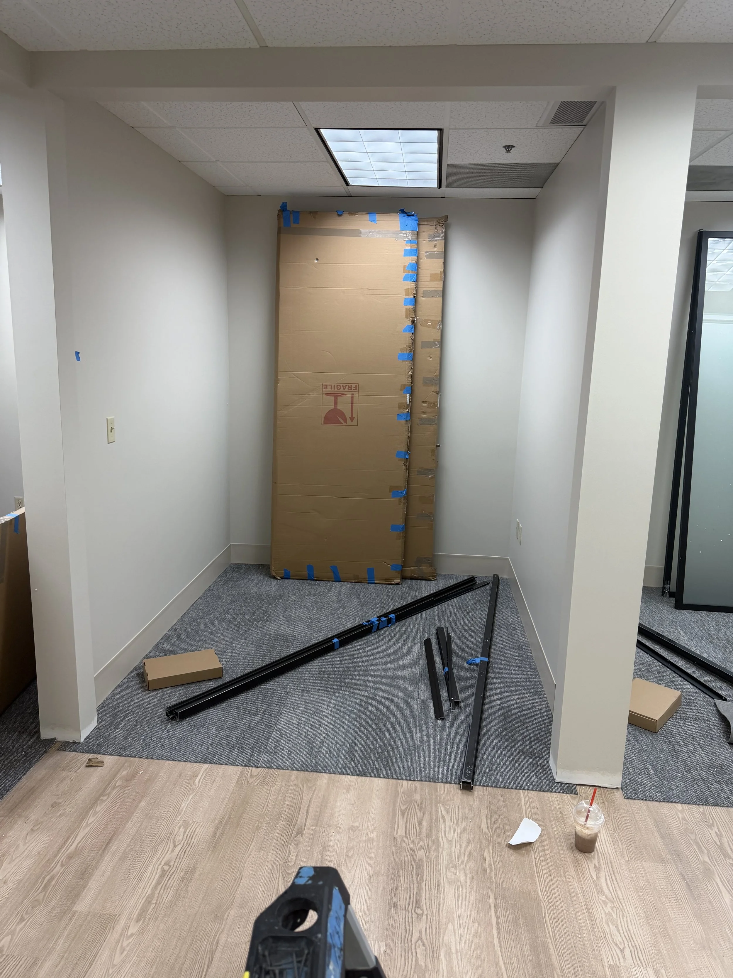 A small office room under construction with a partially assembled black metal frame on the floor, a large cardboard box leaning against the back wall, a cup of iced coffee with a red straw on the right, and a construction tool in the foreground.