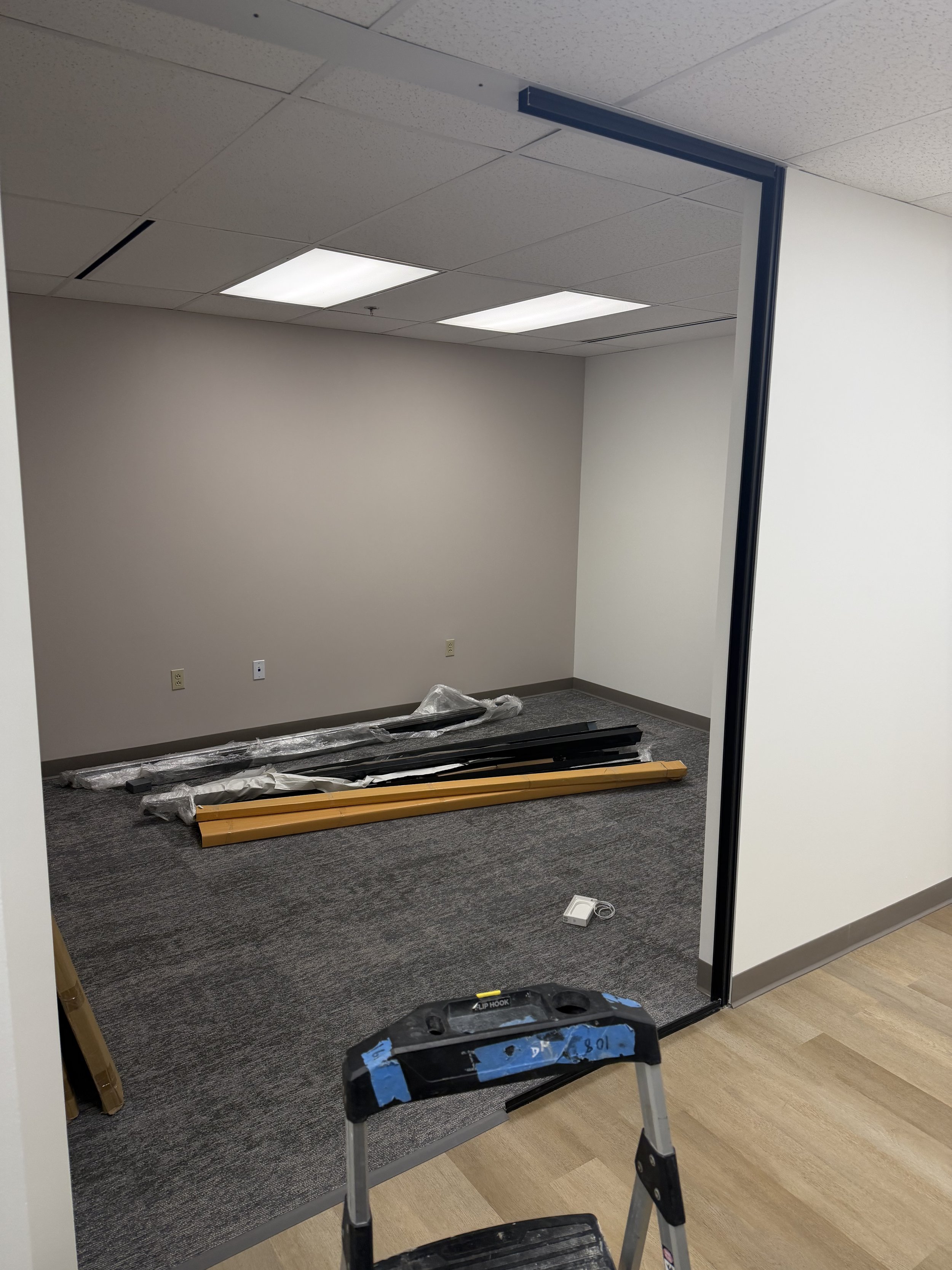 Empty office room with a partially detached wall or partition, construction materials on the carpeted floor, and a small step ladder in the foreground.