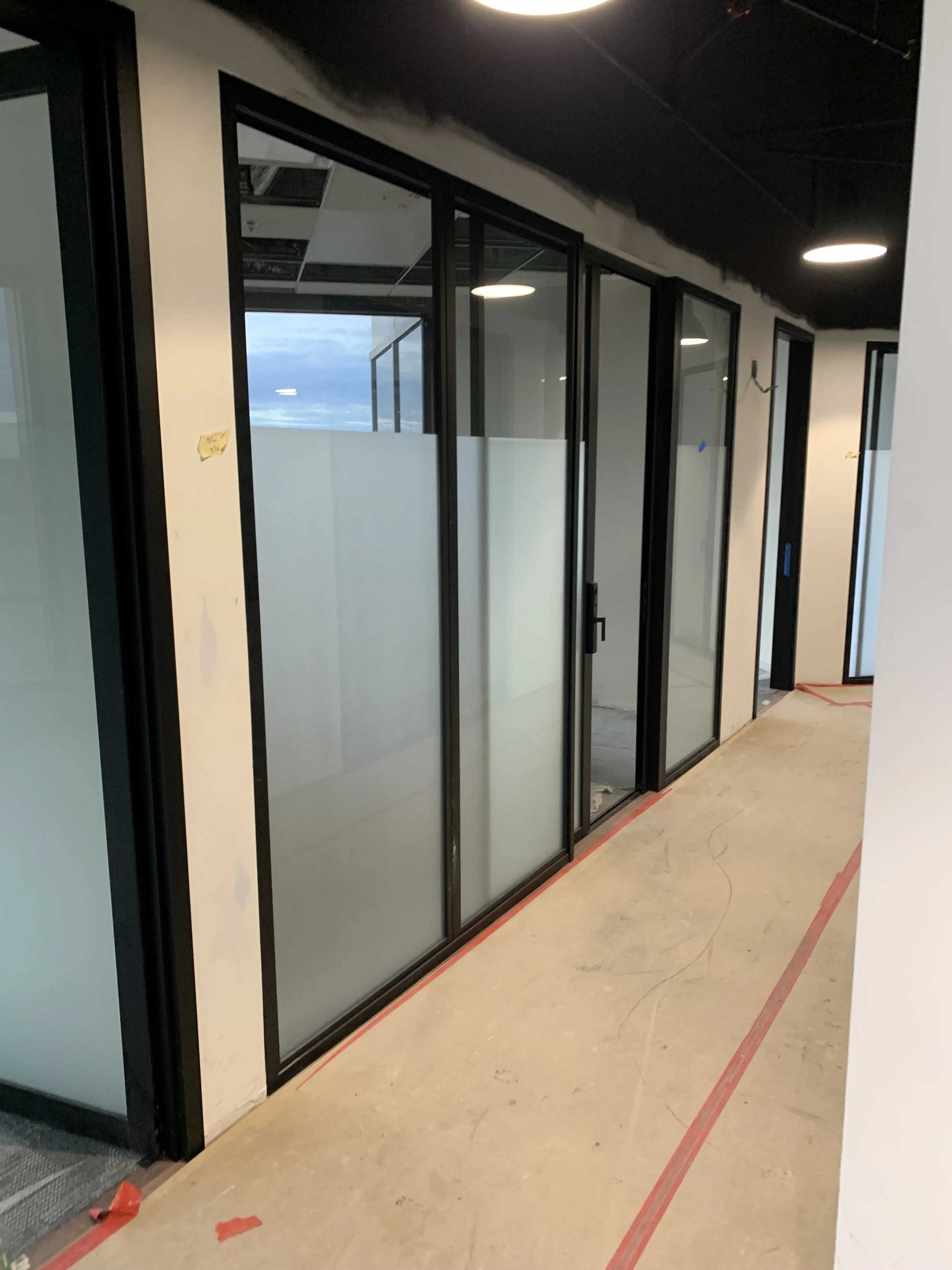 Construction hallway with glass doors and black metal frames, some frosted glass, unfinished floor, and overhead lighting