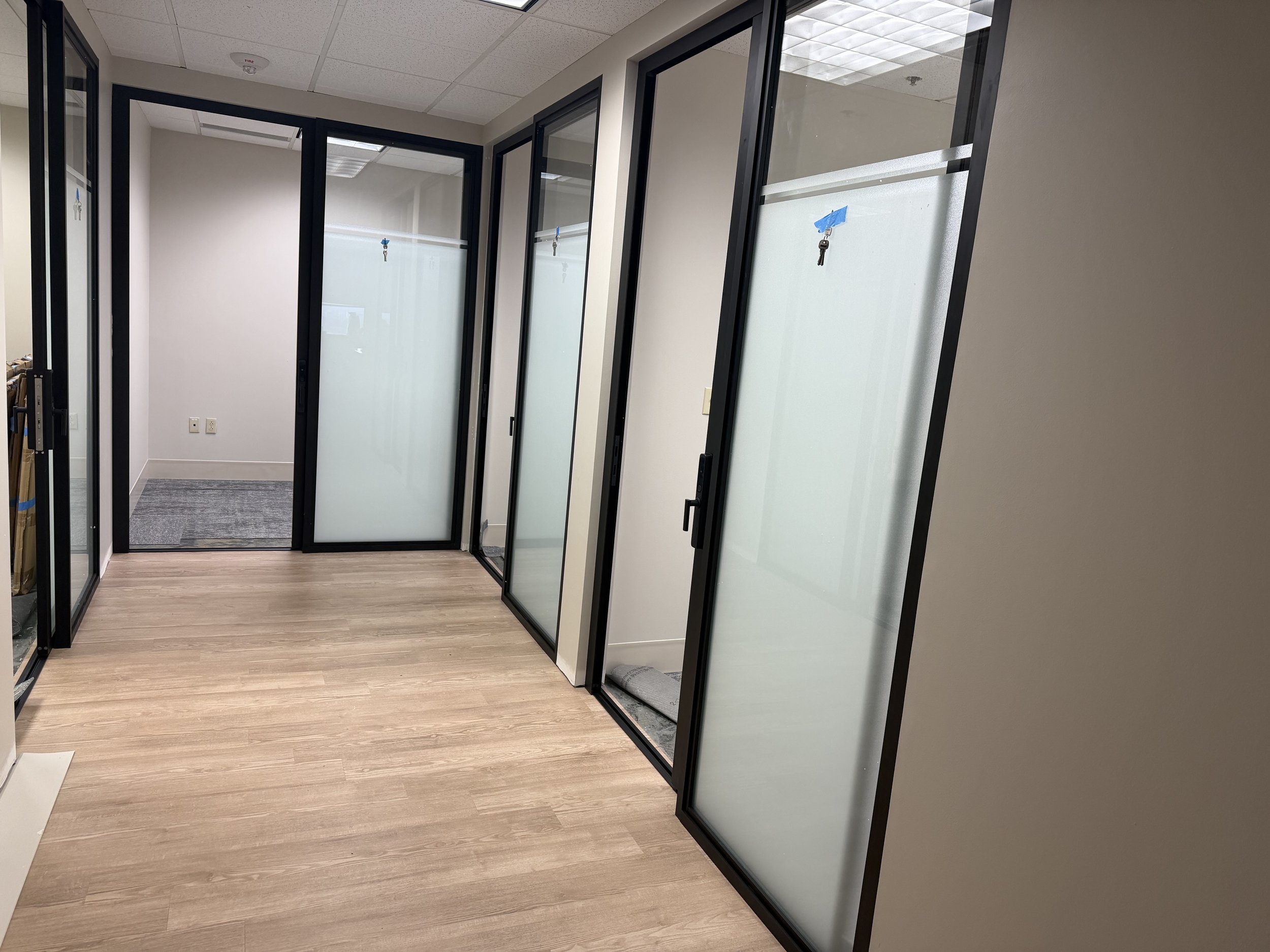 Empty office hallway with glass partitions and frosted glass doors, wood flooring, and ceiling tiles.