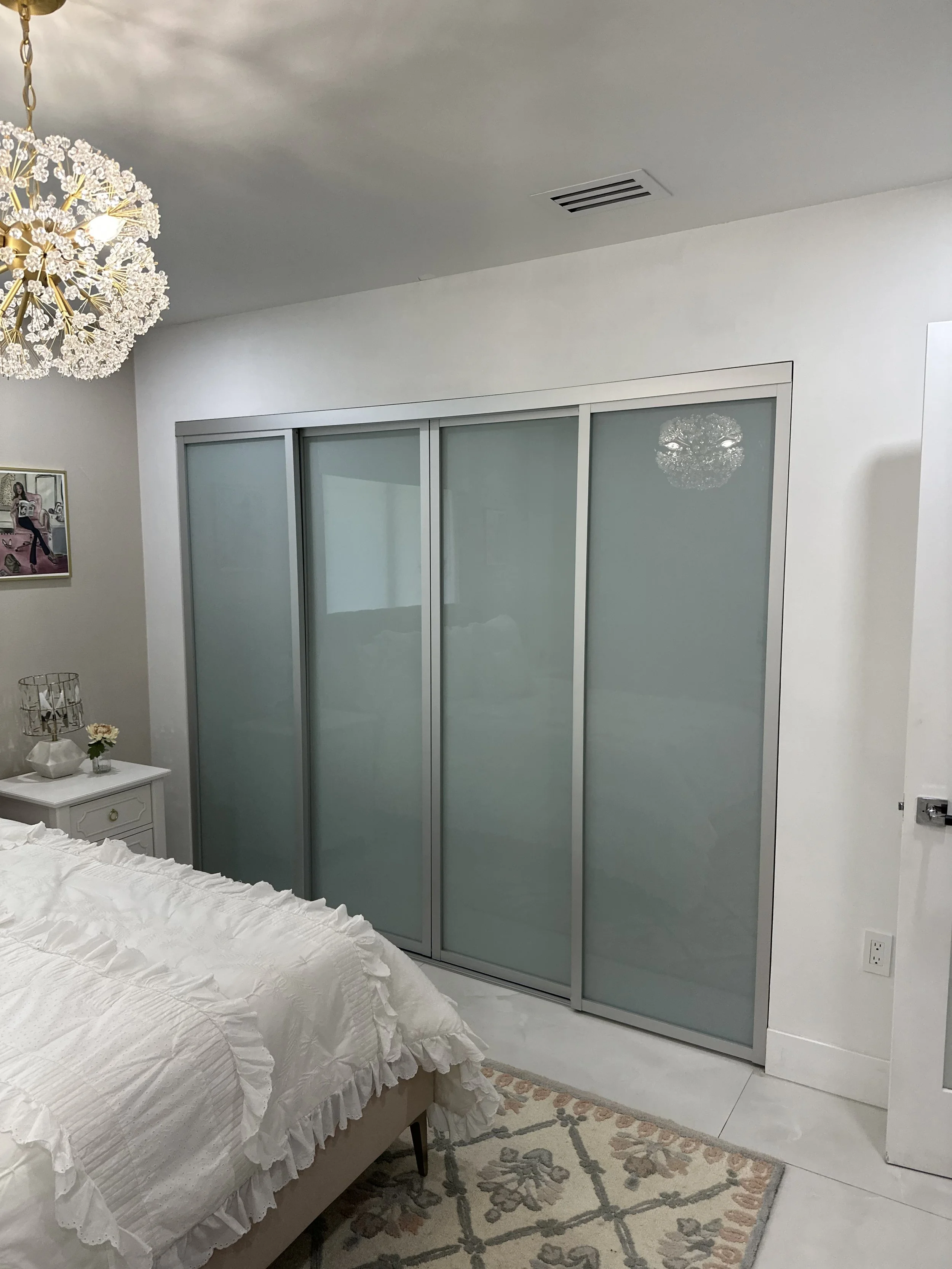A bedroom with a white bedspread, white side table with decorative items, and a sliding frosted glass closet door. There is a floral rug on the floor and a chandelier hanging from the ceiling.