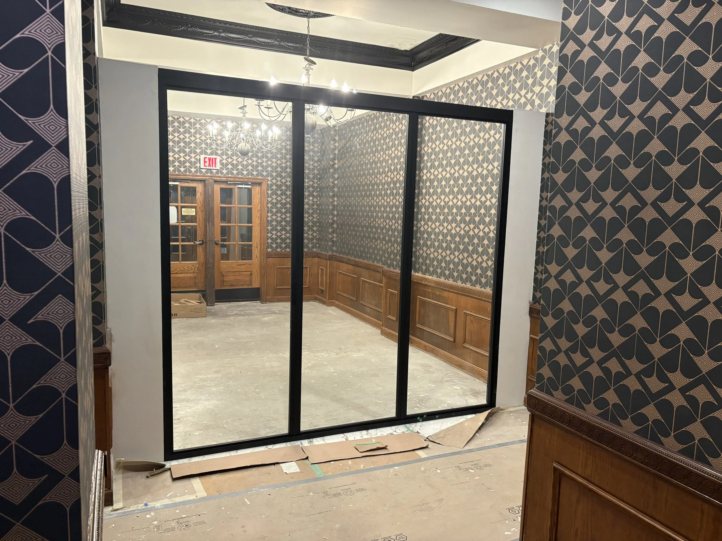 An indoor space under renovation with patterned wallpaper, wooden panels, a chandelier, and a glass partition with black framing.