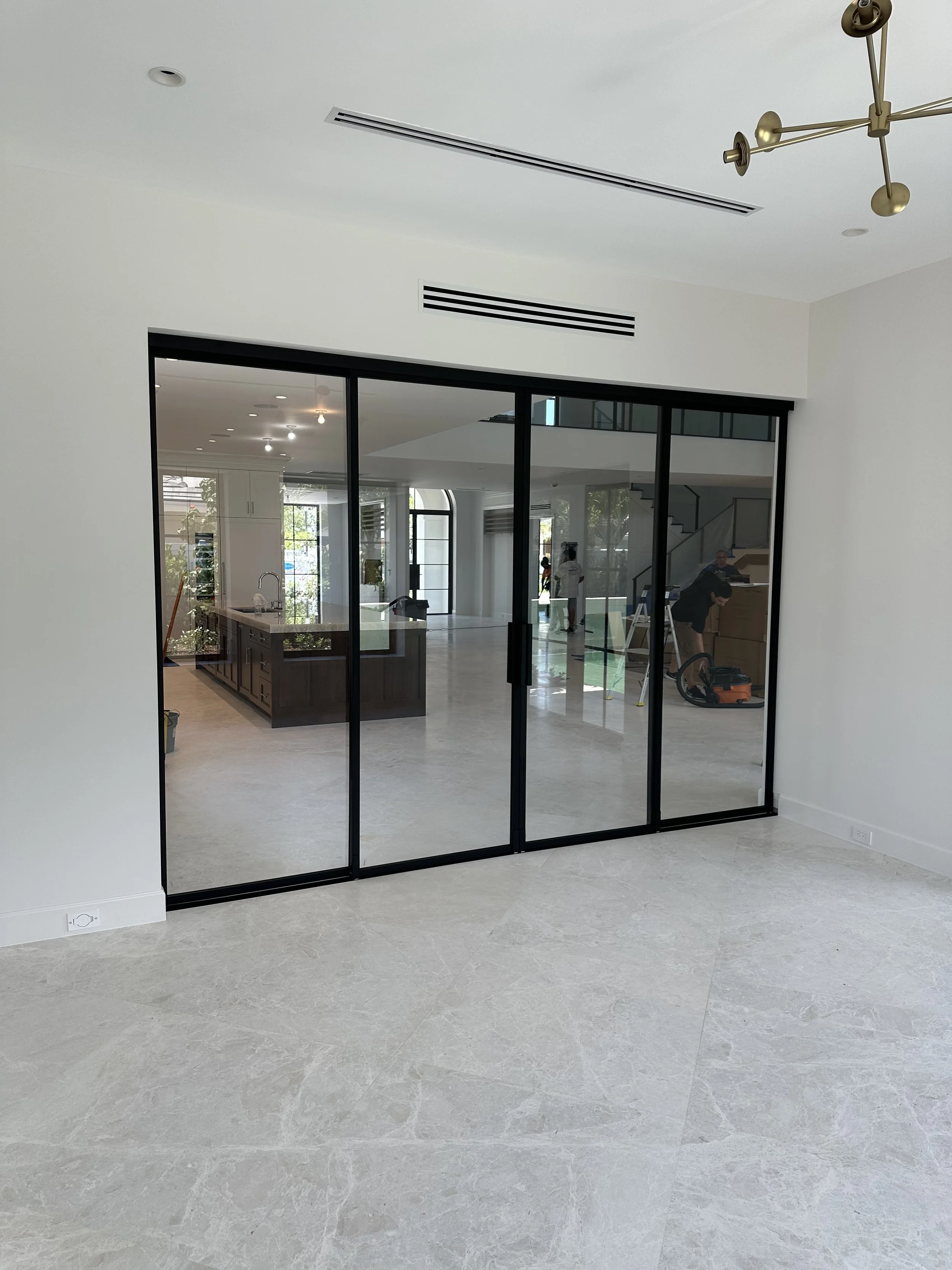 Modern interior space with a black-framed sliding glass door, views into a kitchen and living area, and light-colored flooring.