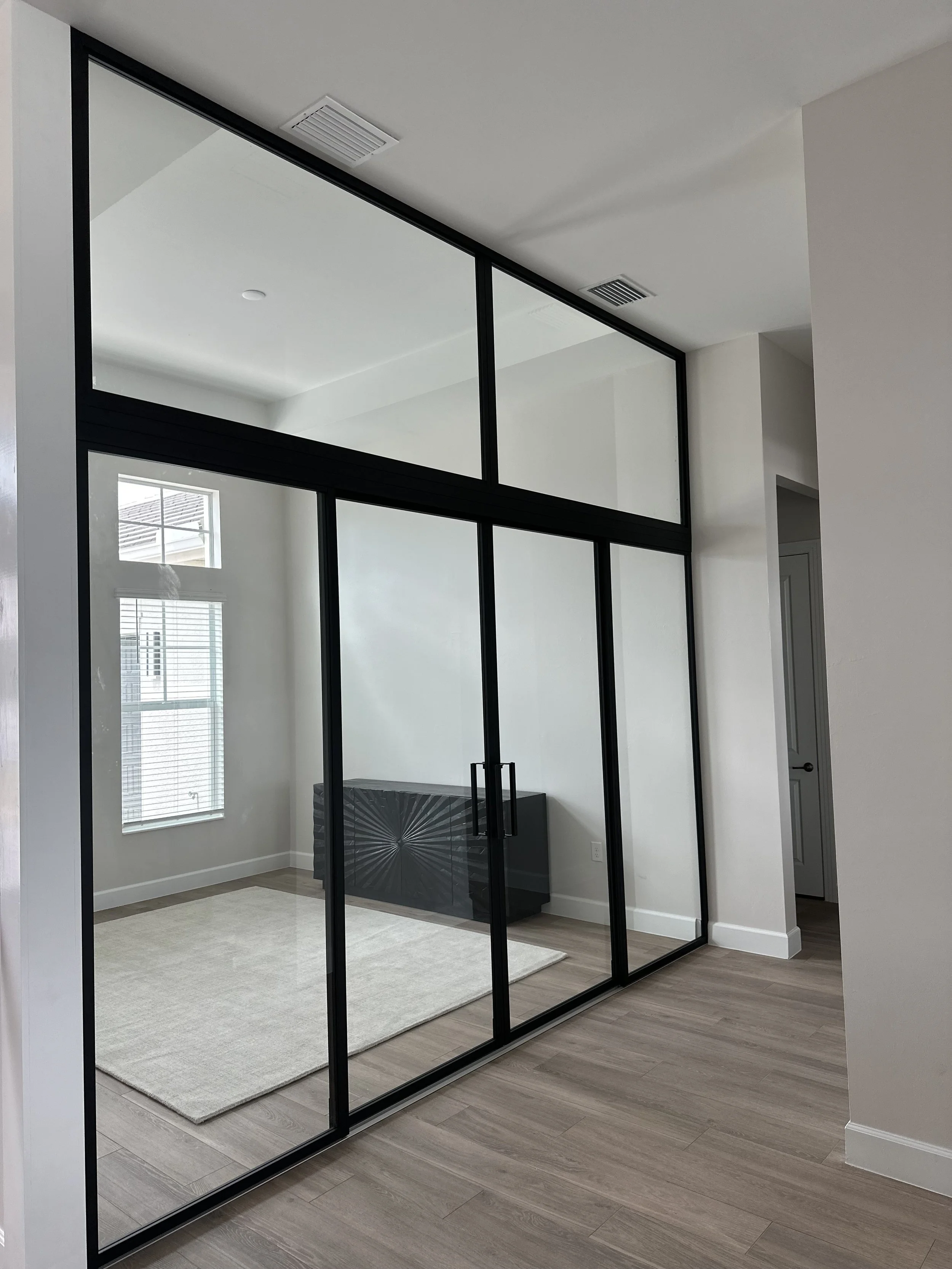 Interior of a modern room with a glass partition, hardwood floors, a window with blinds, a rug, and a black cabinet.