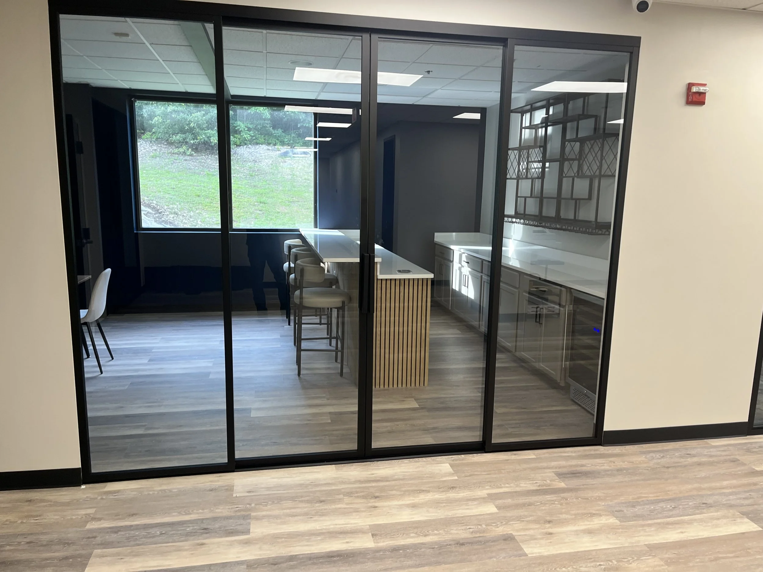 A modern meeting room or lounge area with a glass wall, a window overlooking greenery outside, a high table with beige chairs, white cabinetry, and a decorative black metal shelf against the wall.