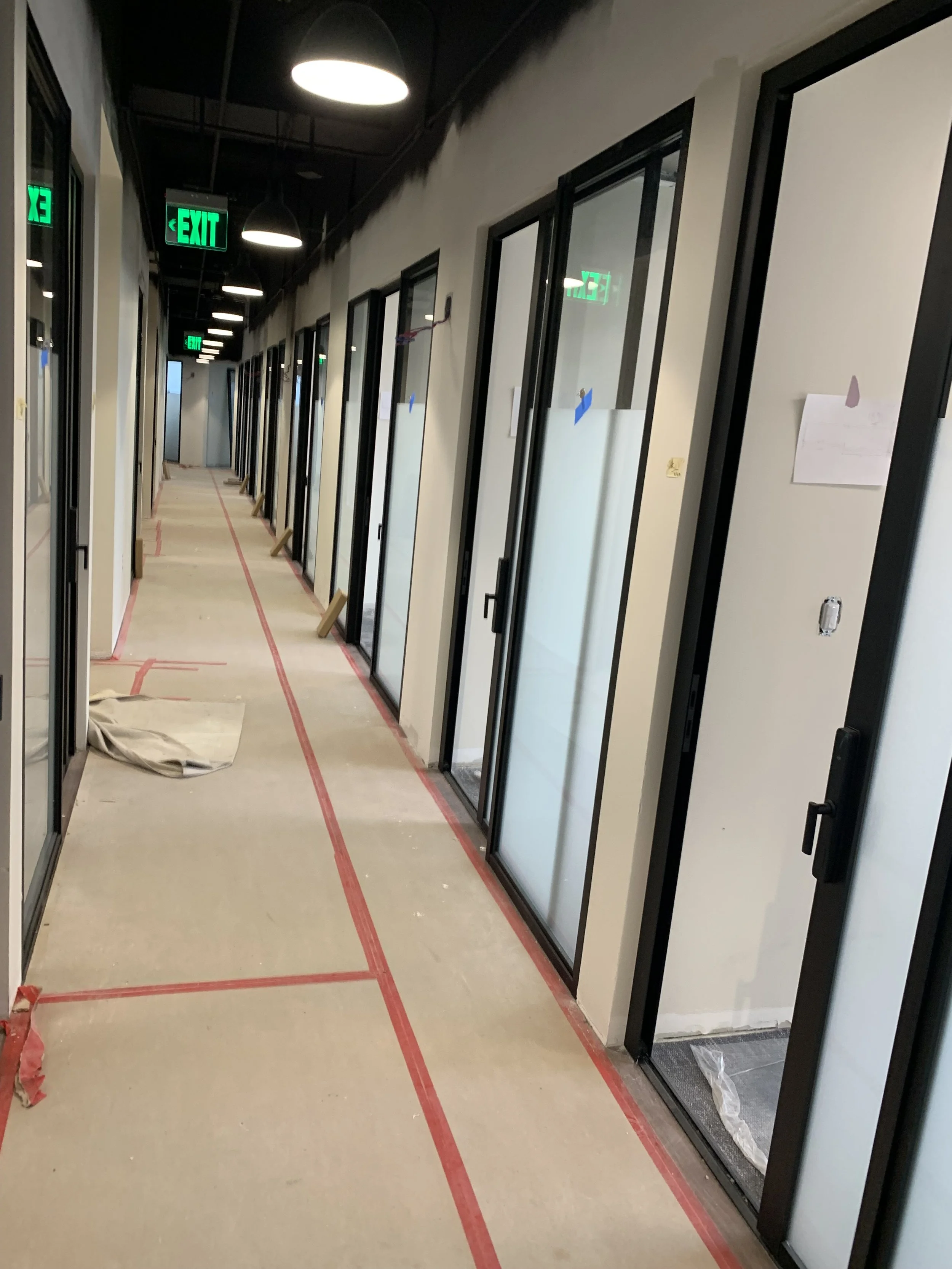 Hallway under construction with closed glass door offices on each side, green exit signs, and protective tape on the floor.