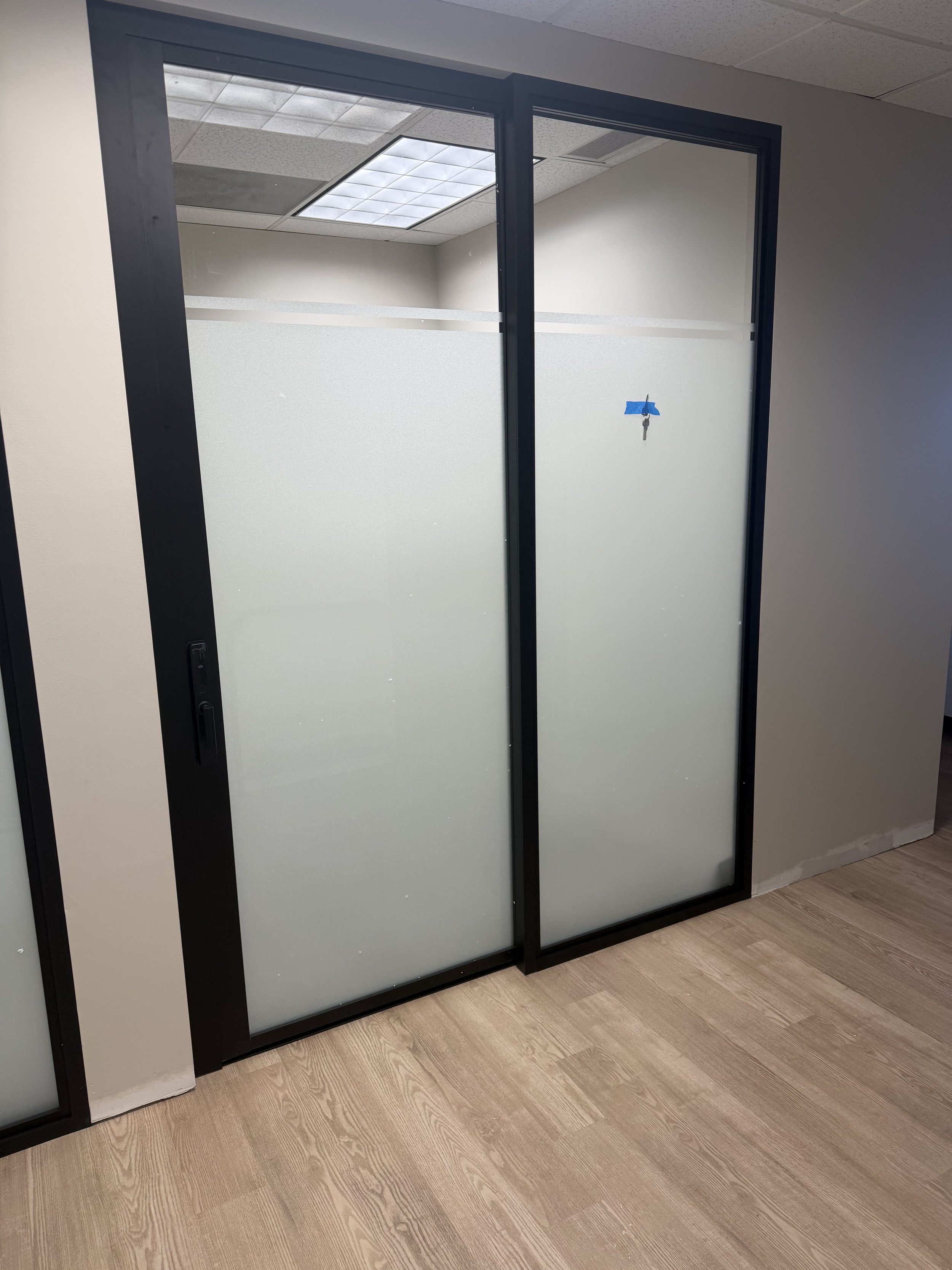 Office glass door with frosted lower half and a lock taped with blue painter's tape, leading to another office space with a ceiling light and drop ceiling tiles.
