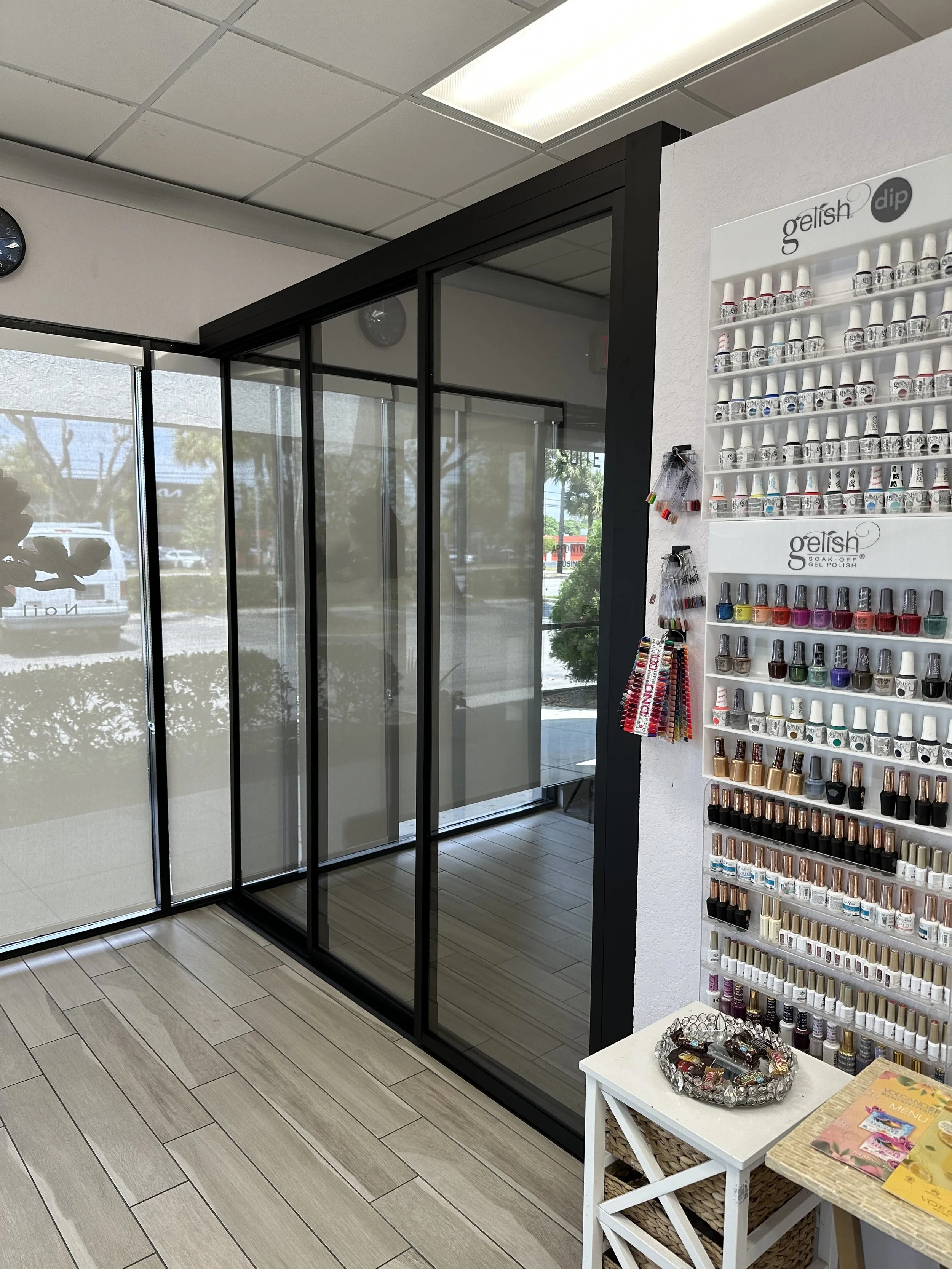A glass display shower enclosure with black framing inside a nail salon, next to a wall-mounted nail polish display with various colors and brands, and a small white table with jewelry inside a store with tiled flooring and large windows.