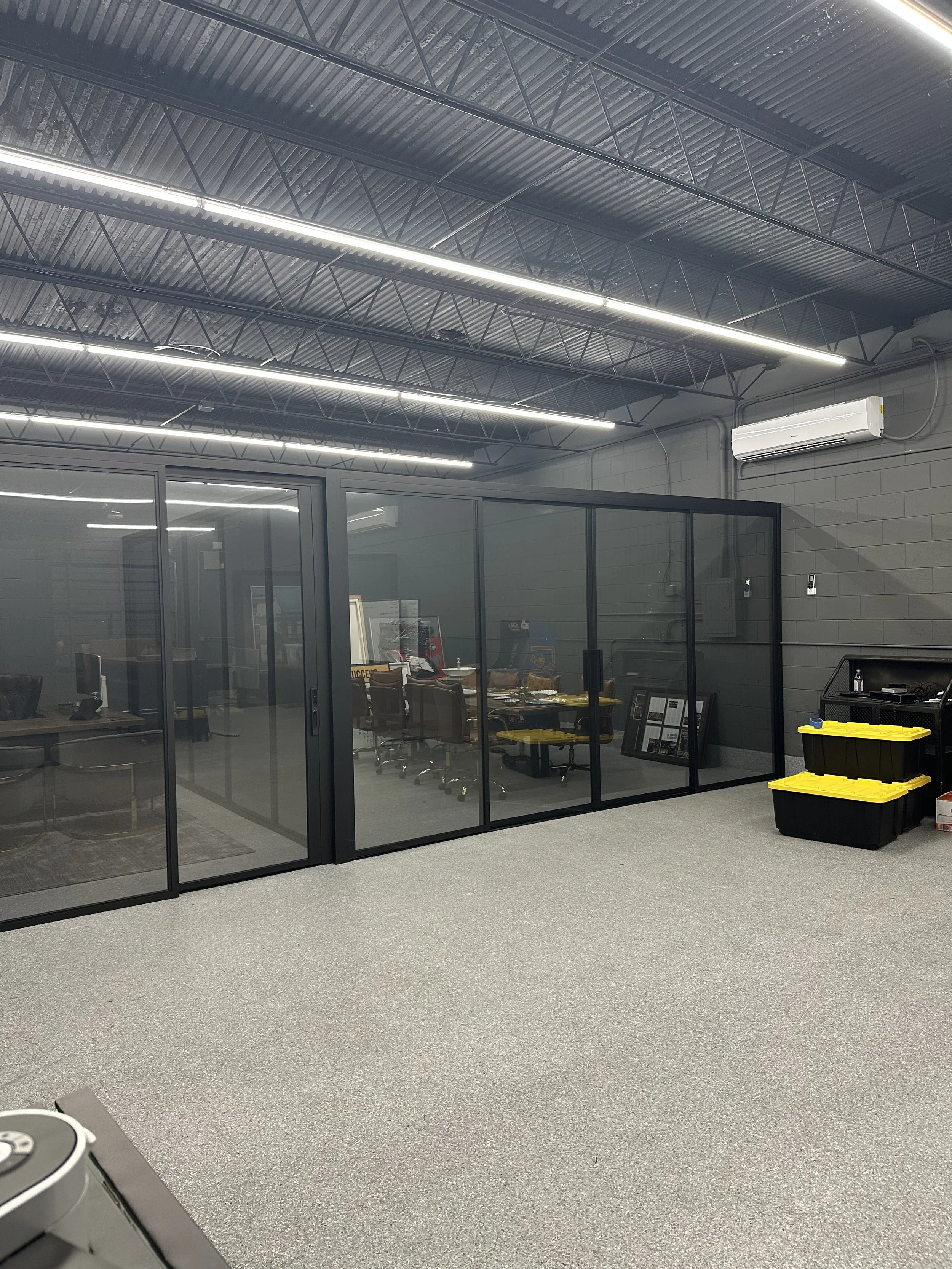 An office conference room with a glass wall, gray walls, and gray carpet, containing a long table with chairs, and a black storage cabinet with yellow storage bins outside the room; the ceiling has exposed metal beams and fluorescent lighting, with a wall-mounted air conditioning unit.