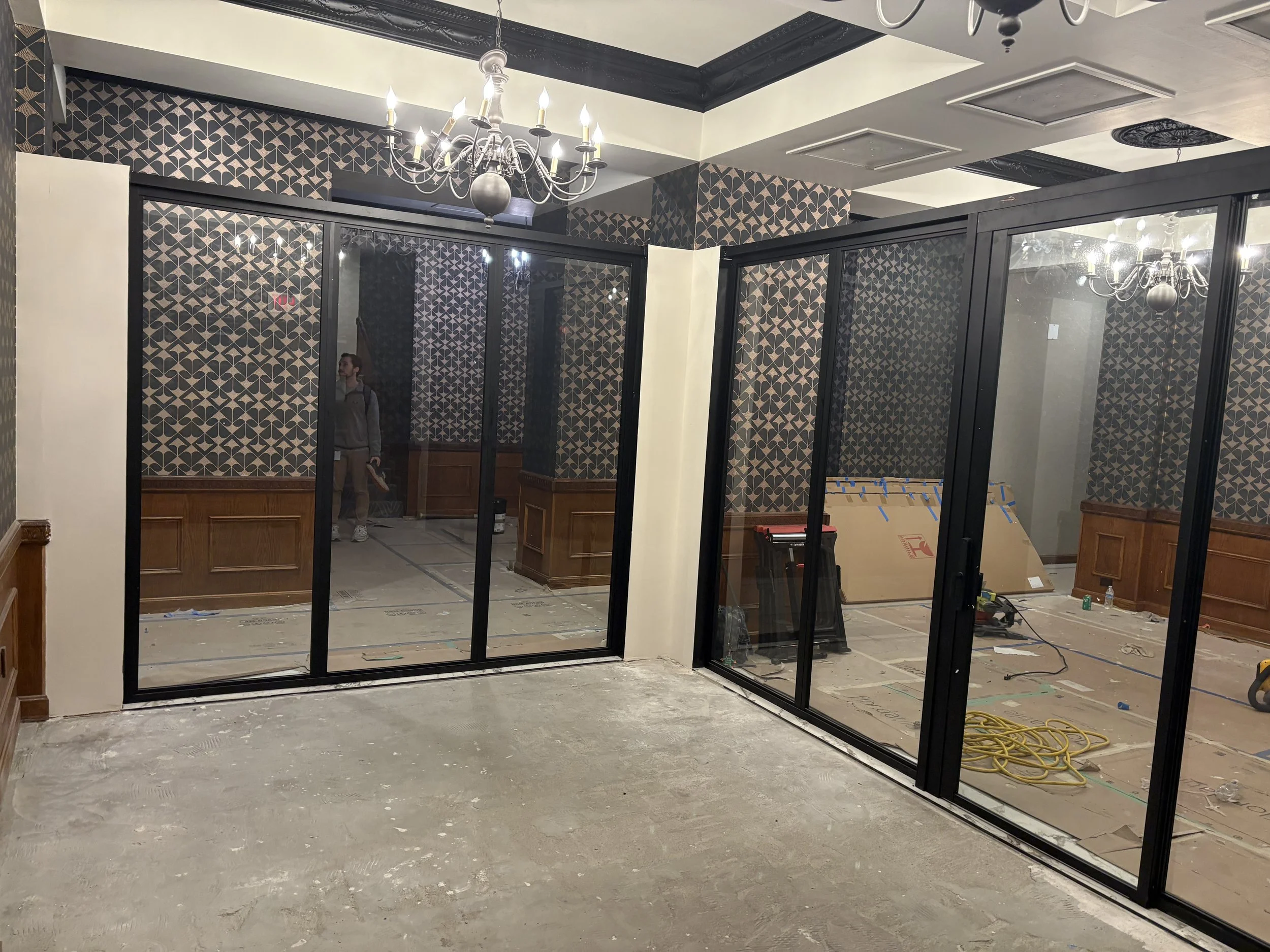 Interior of a room under renovation with glass partition walls, a chandelier, patterned wallpaper, and construction tools and materials on the floor.