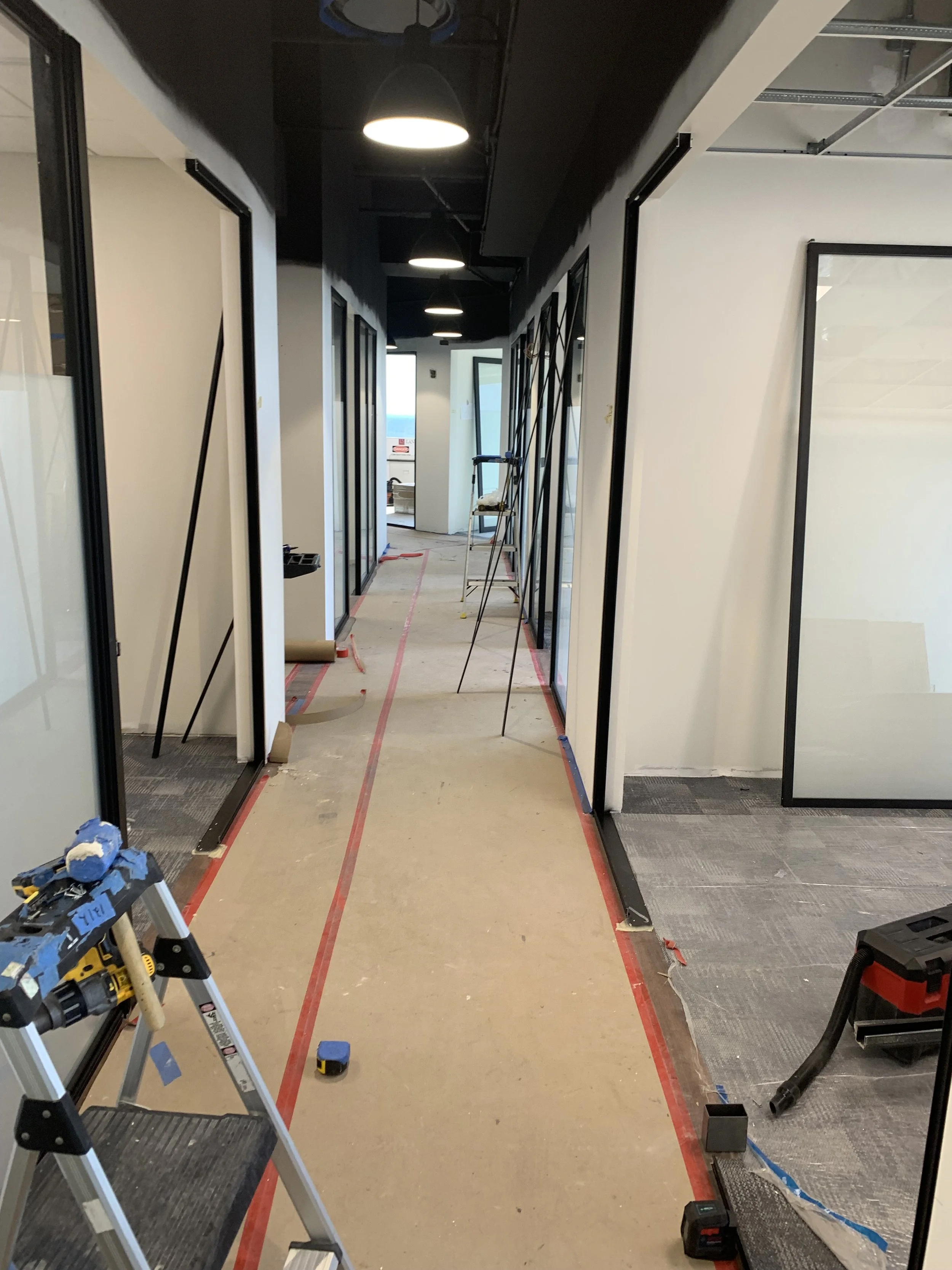 Hallway under construction with tools and ladders, glass wall doors on both sides, and recessed lighting.