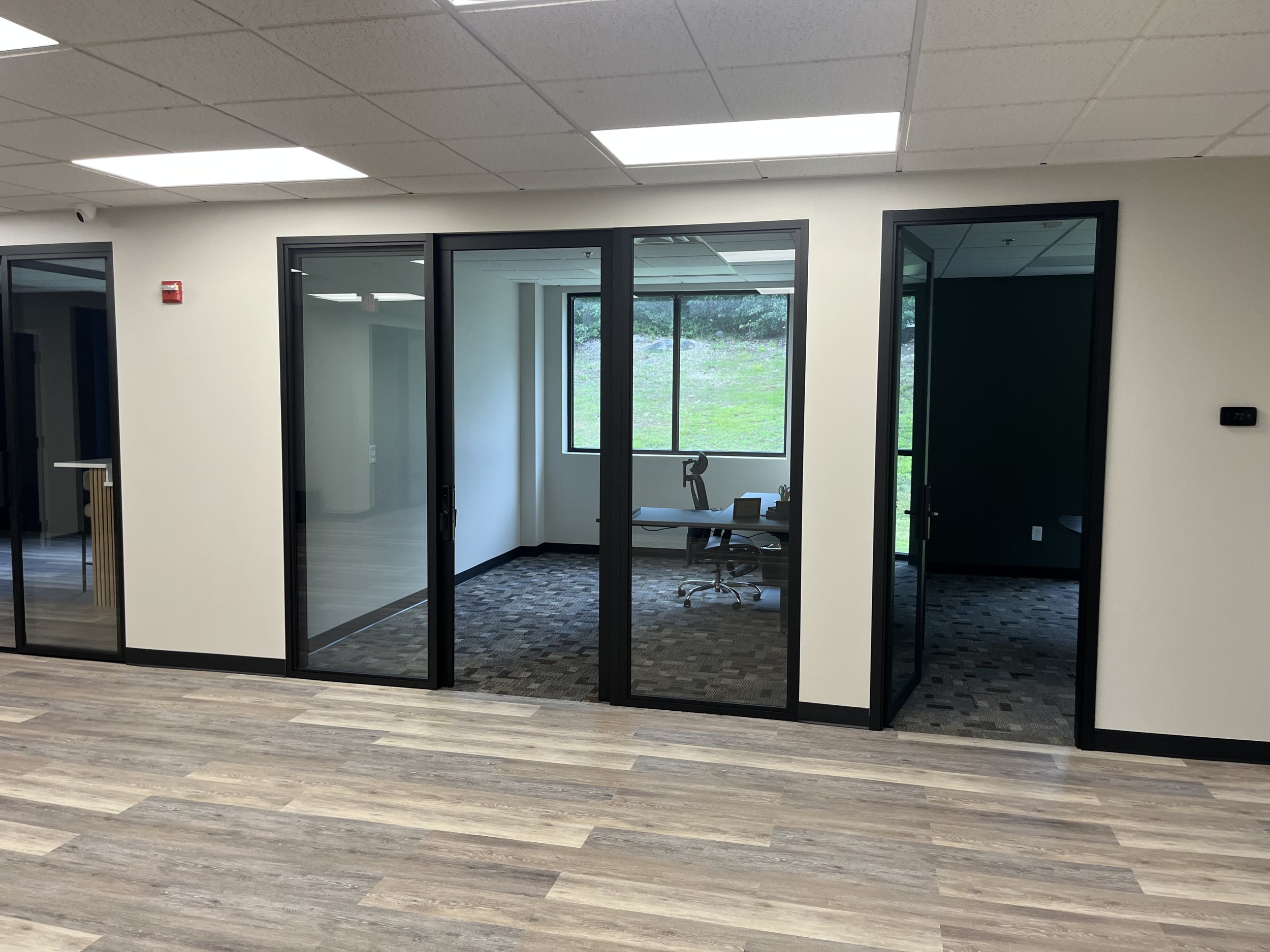 Office space with glass enclosed rooms, view through windows showing greenery outside, and hardwood flooring.