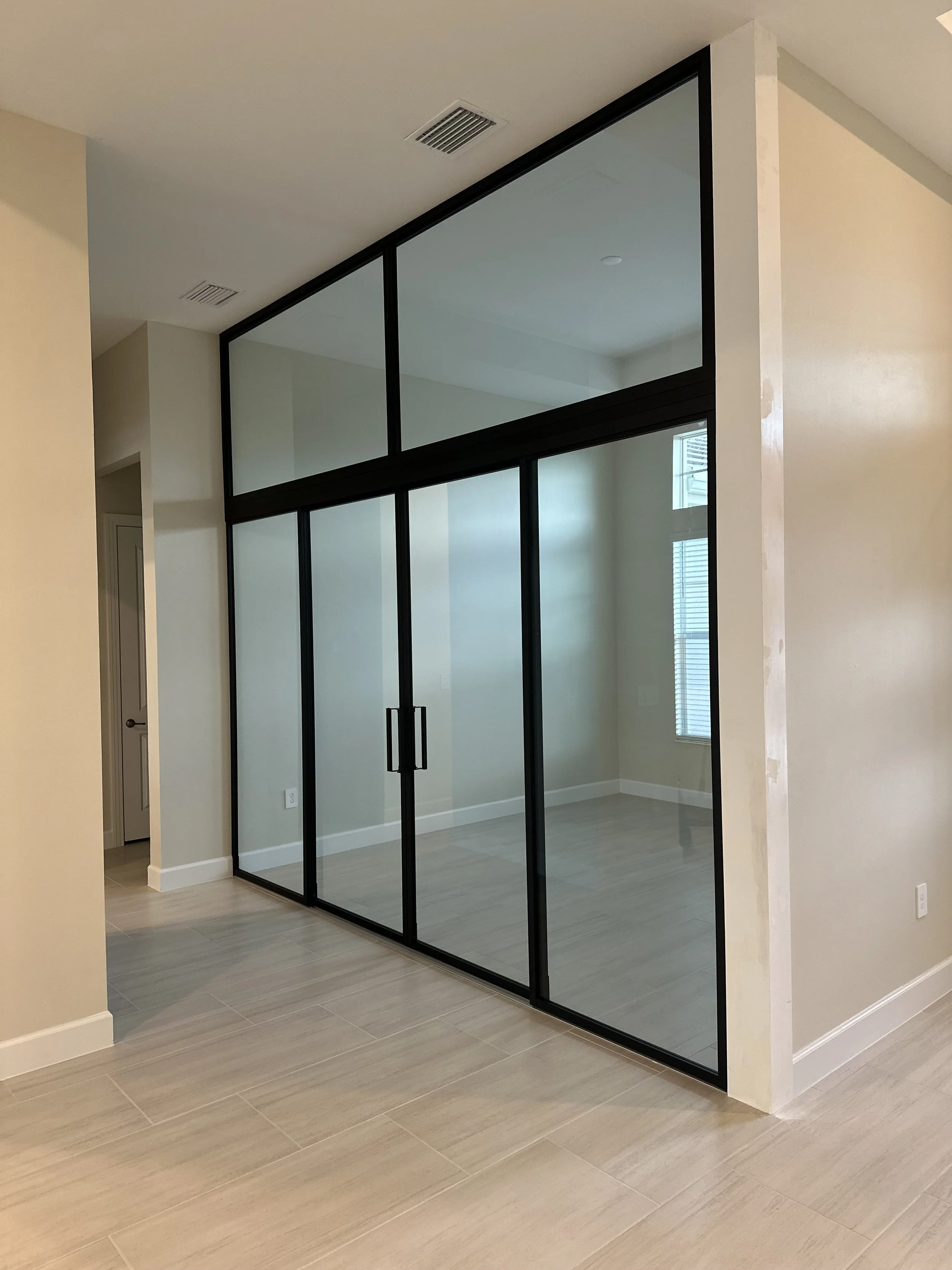 A modern indoor space with a glass partition framed in black, dividing two areas of the room. The floor is covered with light-colored tiles, and there are windows with blinds visible on the far wall.