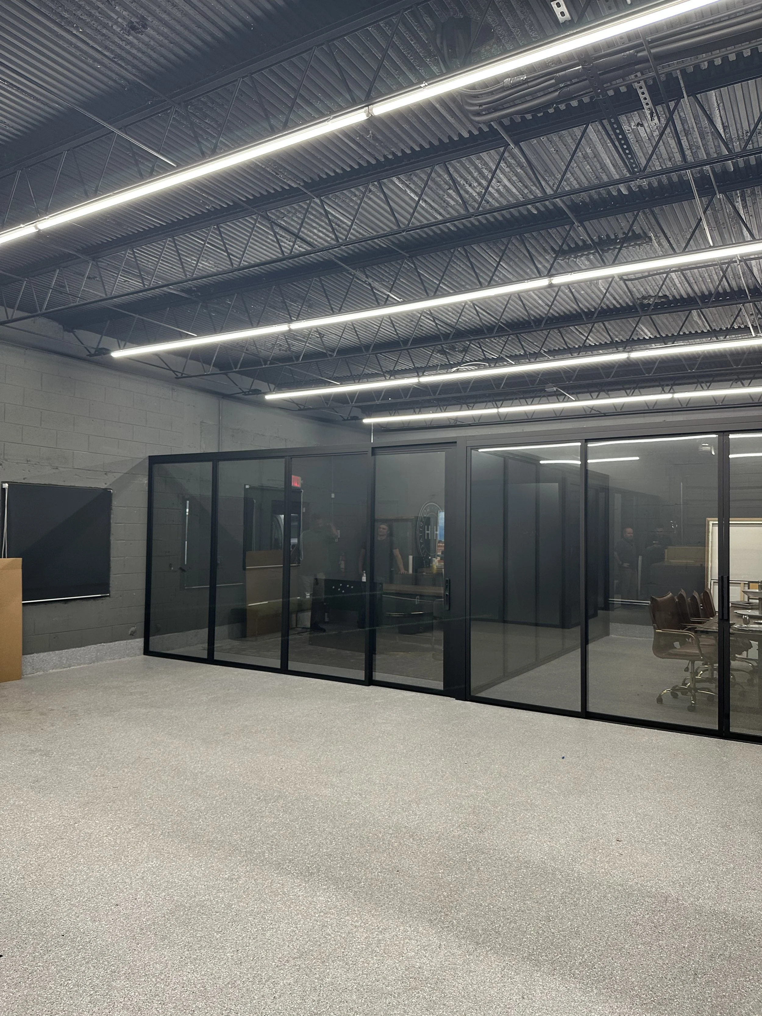 An office conference room with glass walls and a table surrounded by chairs, located in an industrial-style space with exposed beam ceilings and fluorescent lighting.