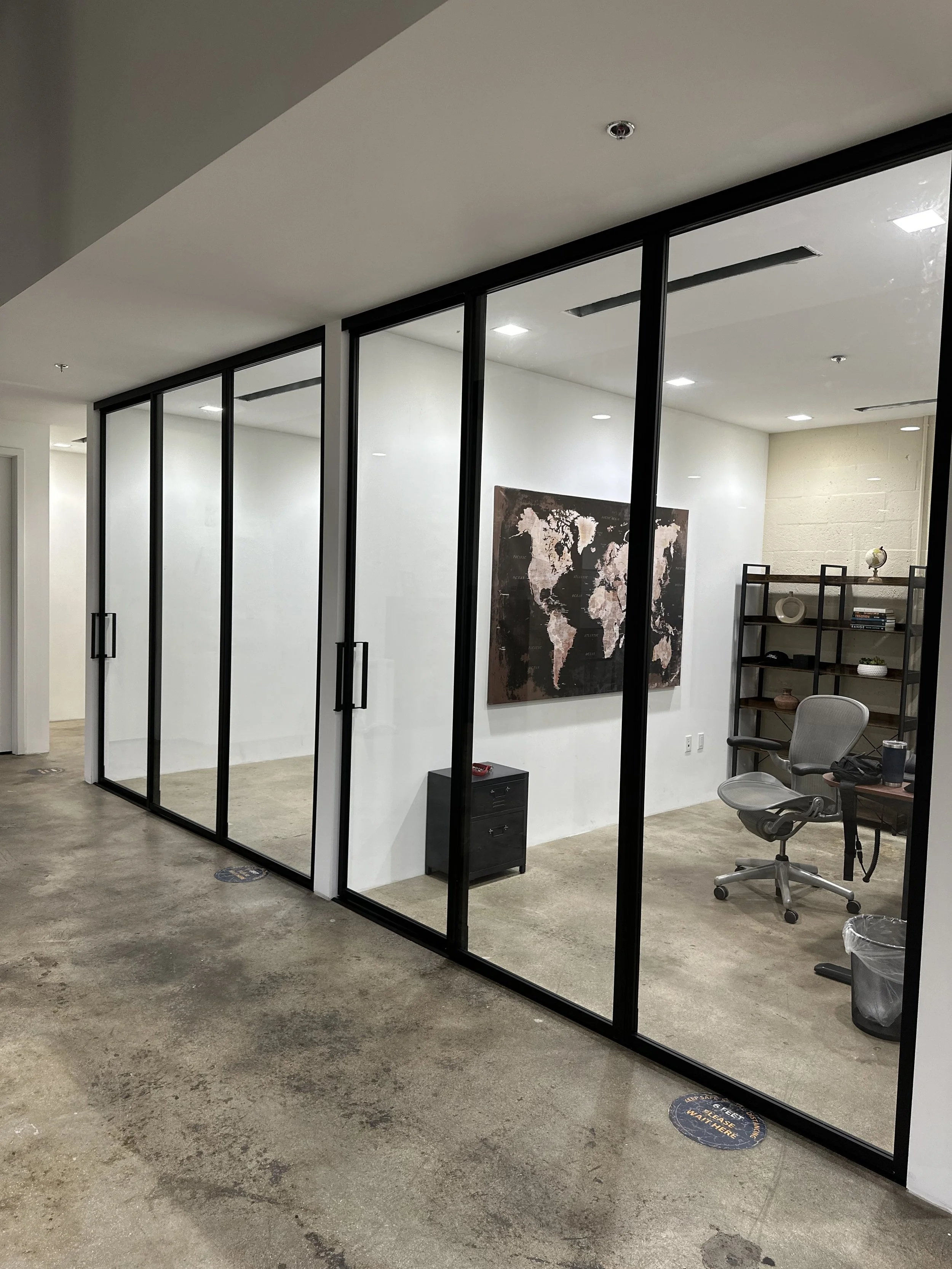 An office with glass walls, a world map on the wall, a bookshelf with decorative items, a rolling office chair, and a trash can.