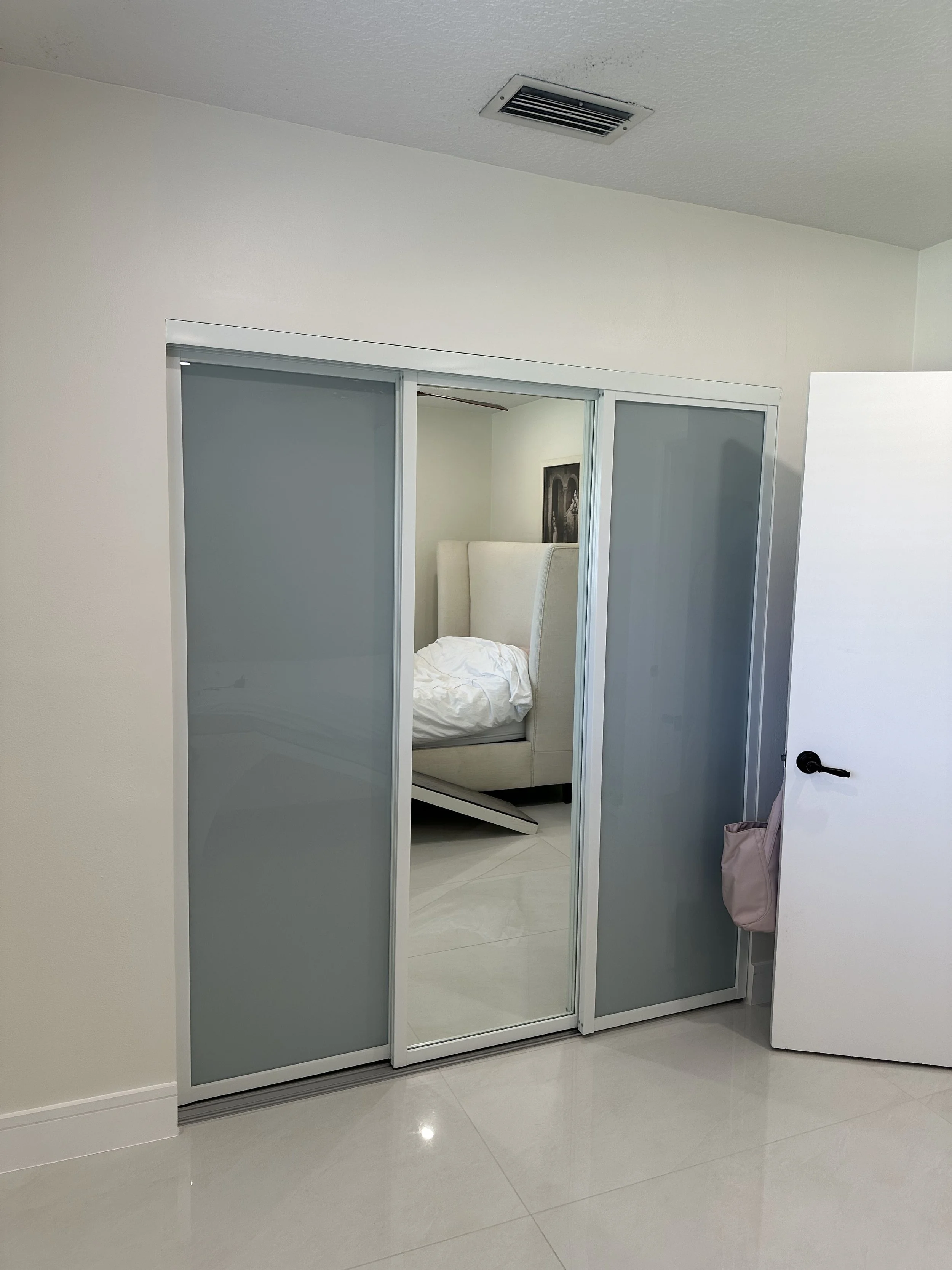 Room with frosted glass sliding closet doors, a white door partially open to the right, and a bed with a white duvet inside the closet room.