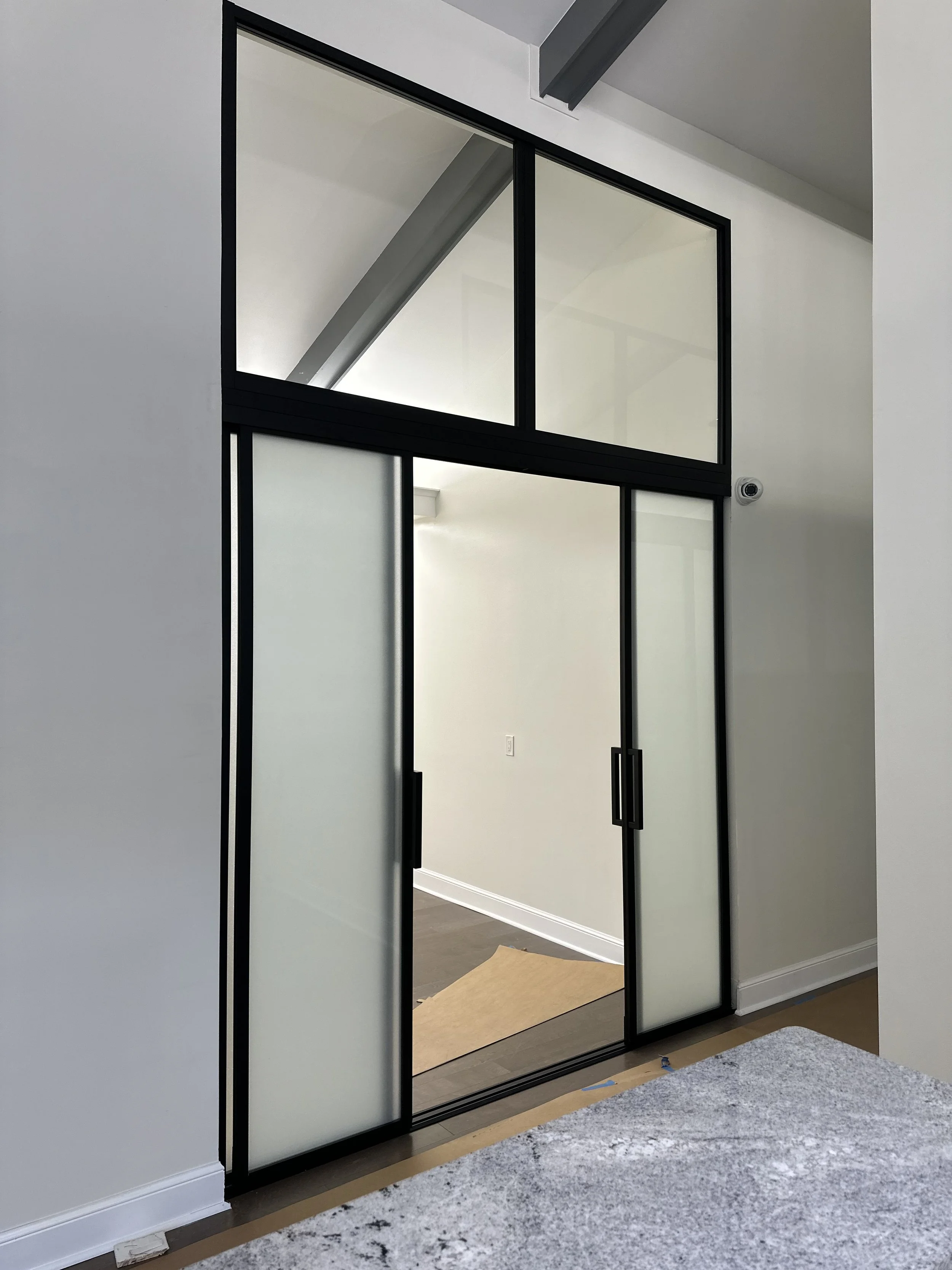 Sliding glass door with black frame separates a room from the hallway. The glass is frosted, and there is a high window above the door. A security camera is mounted on the wall near the ceiling.