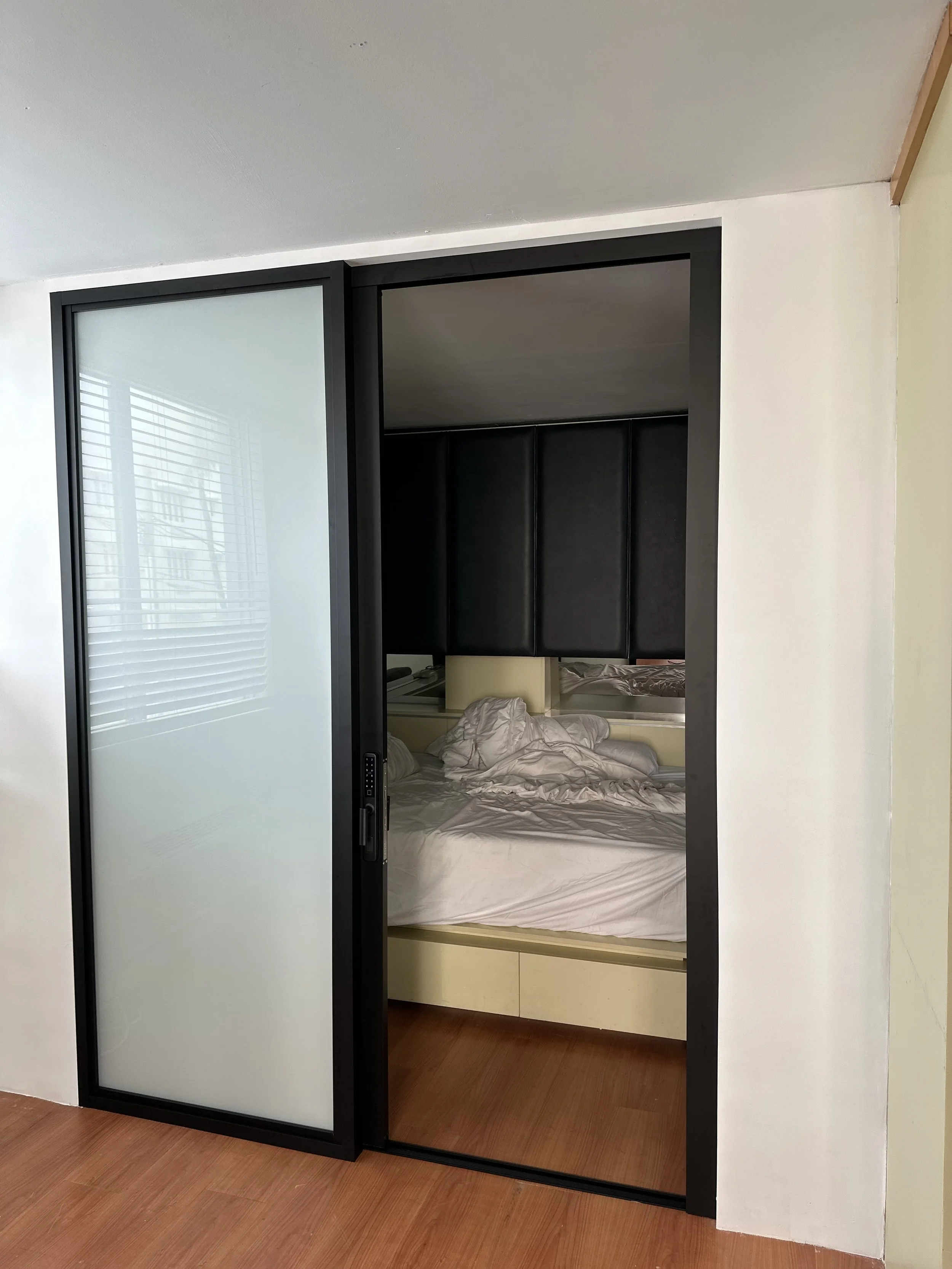 A bedroom viewed through a partially opened sliding frosted glass door with a black frame. Inside, a bed with white sheets and a black padded headboard is visible, and a window with blinds is reflected in the glass door.