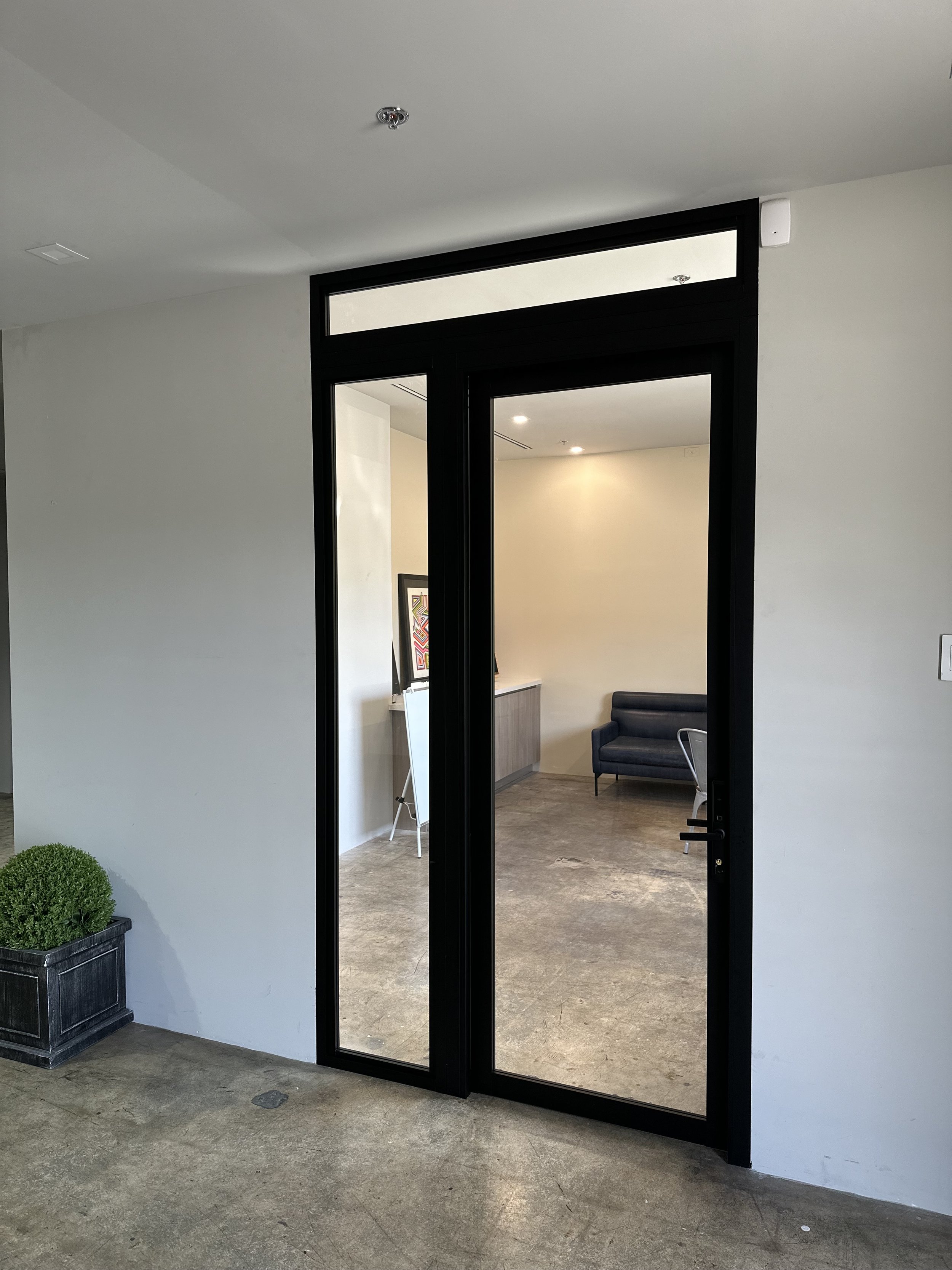 Interior view of an office with a glass door, a black sofa, a framed picture on the wall, and a potted plant outside the door.