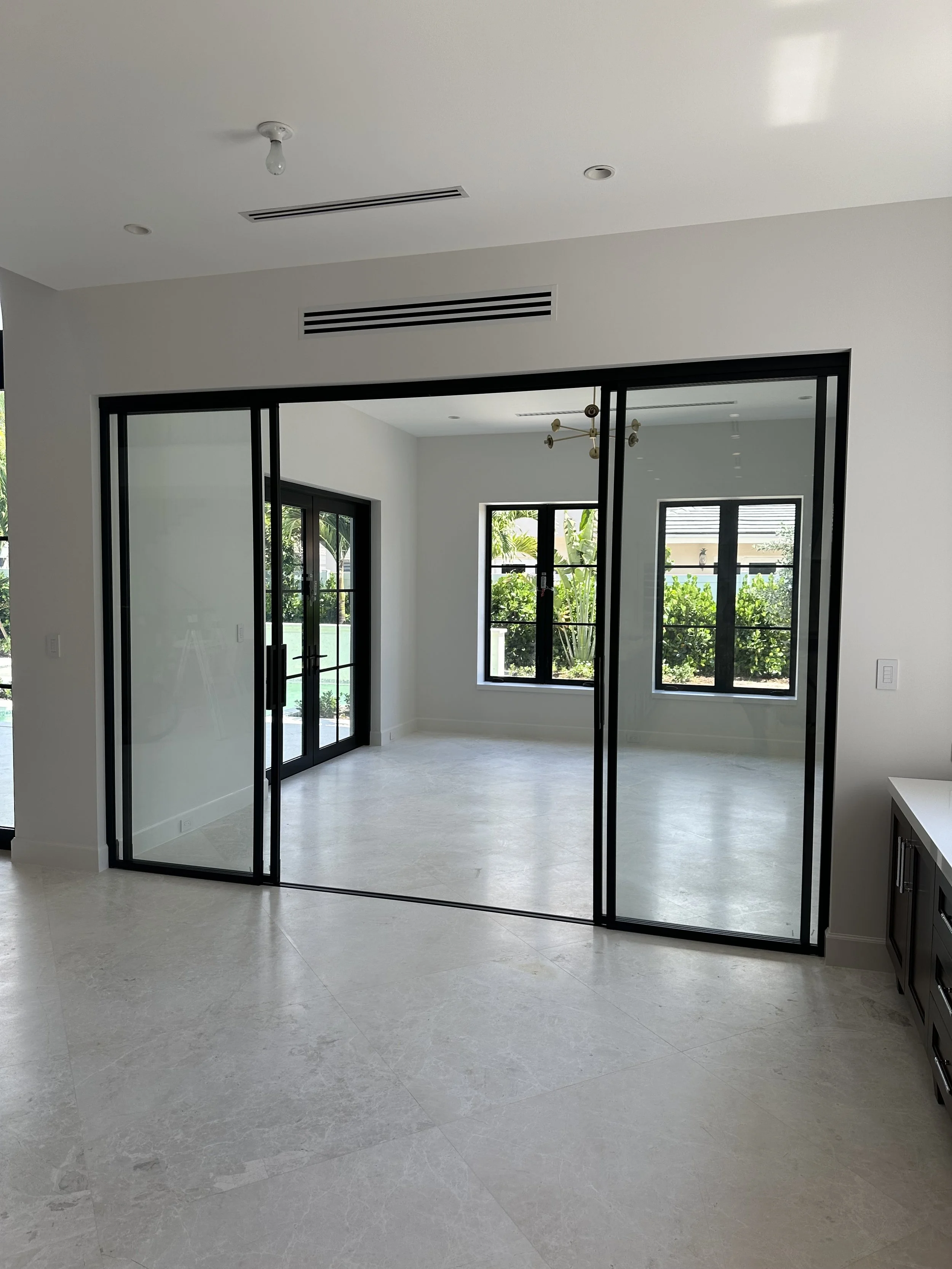 Modern interior space with sliding glass doors, large windows showing greenery outside, white walls, and a minimalistic ceiling light fixture.