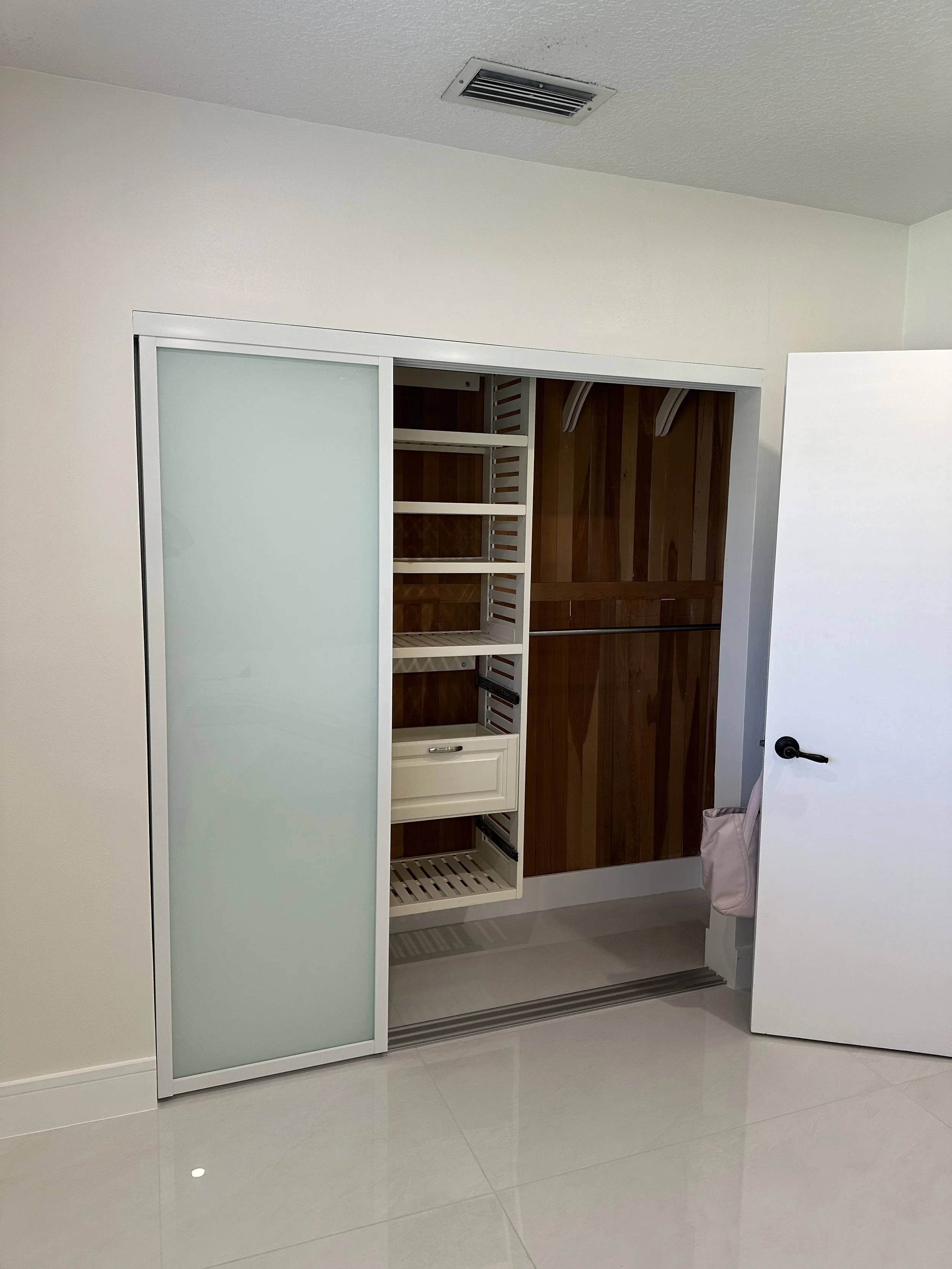 Empty walk-in closet with wooden interior and sliding frosted glass door, shelf and hanging rod, partially open door, and tiled floor.