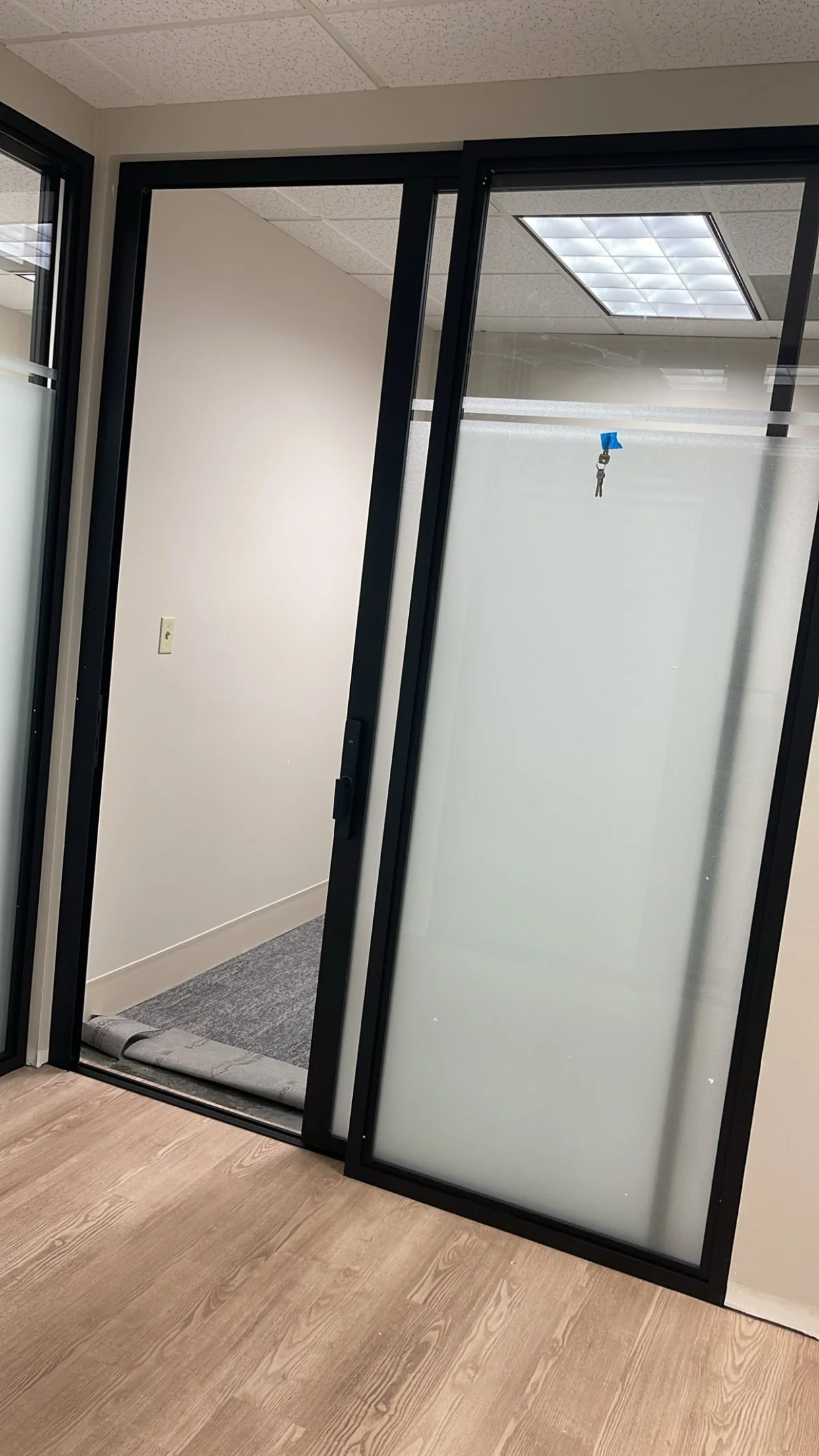 An office with a partially open glass door with frosted glass and black frame, revealing a small room or closet with white walls, gray carpet, a rolled-up mat on the floor, and an overhead ceiling light.