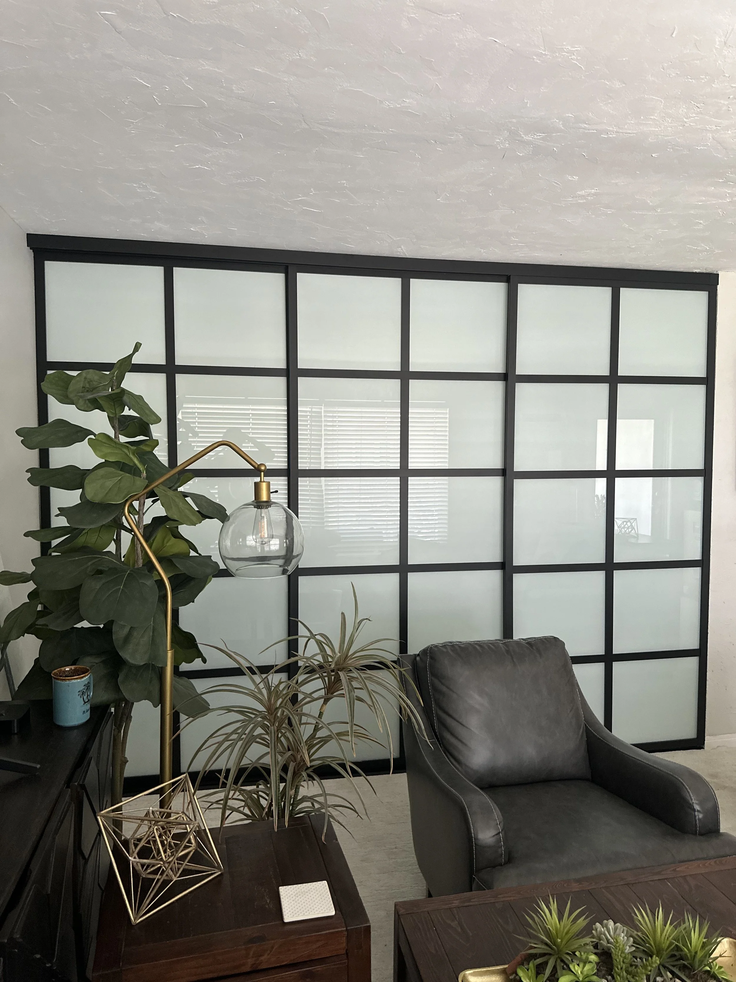 A living room with a black leather armchair, green plants, a modern gold and glass floor lamp, and a large frosted glass panel window with black grid framing.