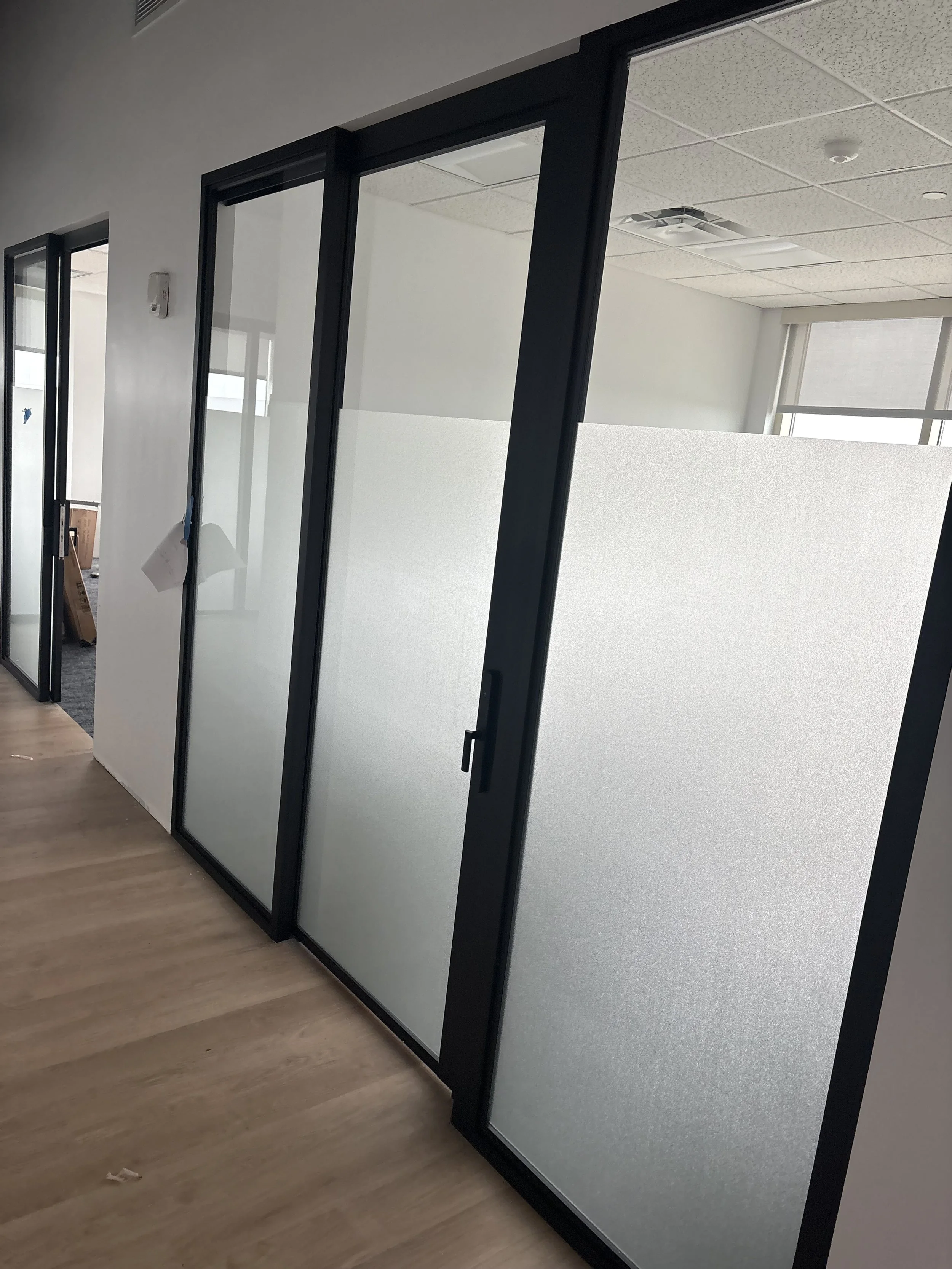 Office hallway with glass partition doors, some frosted and some clear, wood flooring, ceiling panels, and ceiling-mounted light fixtures.