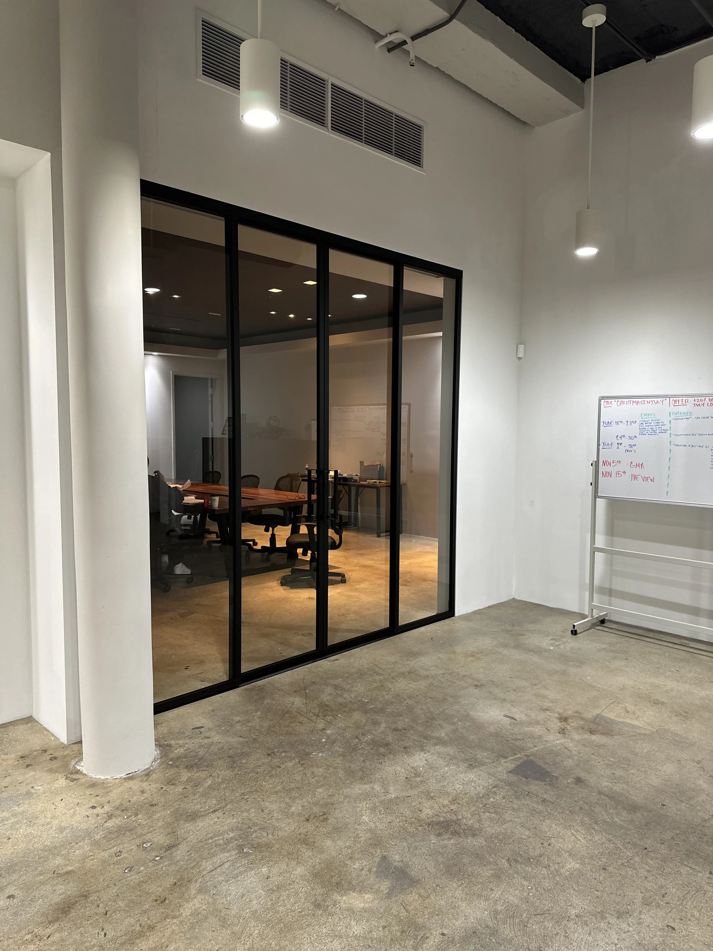An empty room with a concrete floor, white walls, and a glass wall with black frames that shows a conference room with chairs and a table. There are two white pendant lights hanging from the ceiling and a whiteboard on the right side.
