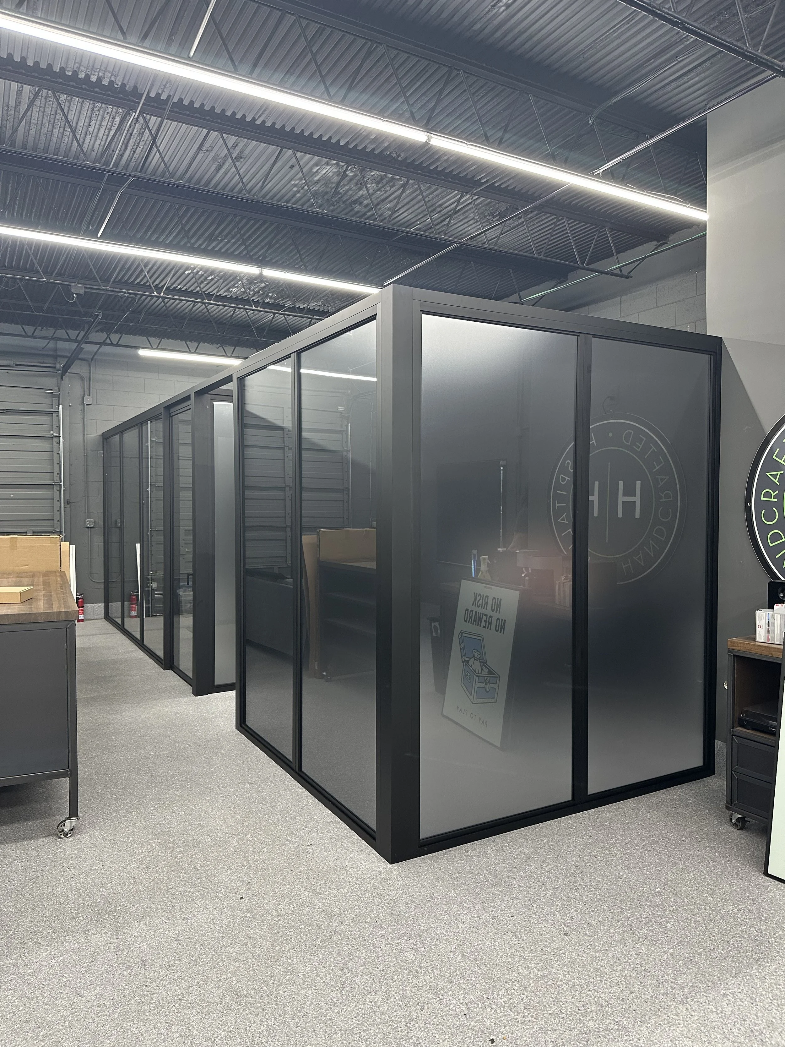 Indoor recreational or gym facility with black-framed glass-enclosed training pods, some containing couches, and a sign inside the front pod that says 'NO REWIND, NO DRAG, PAY TO PLAY'.