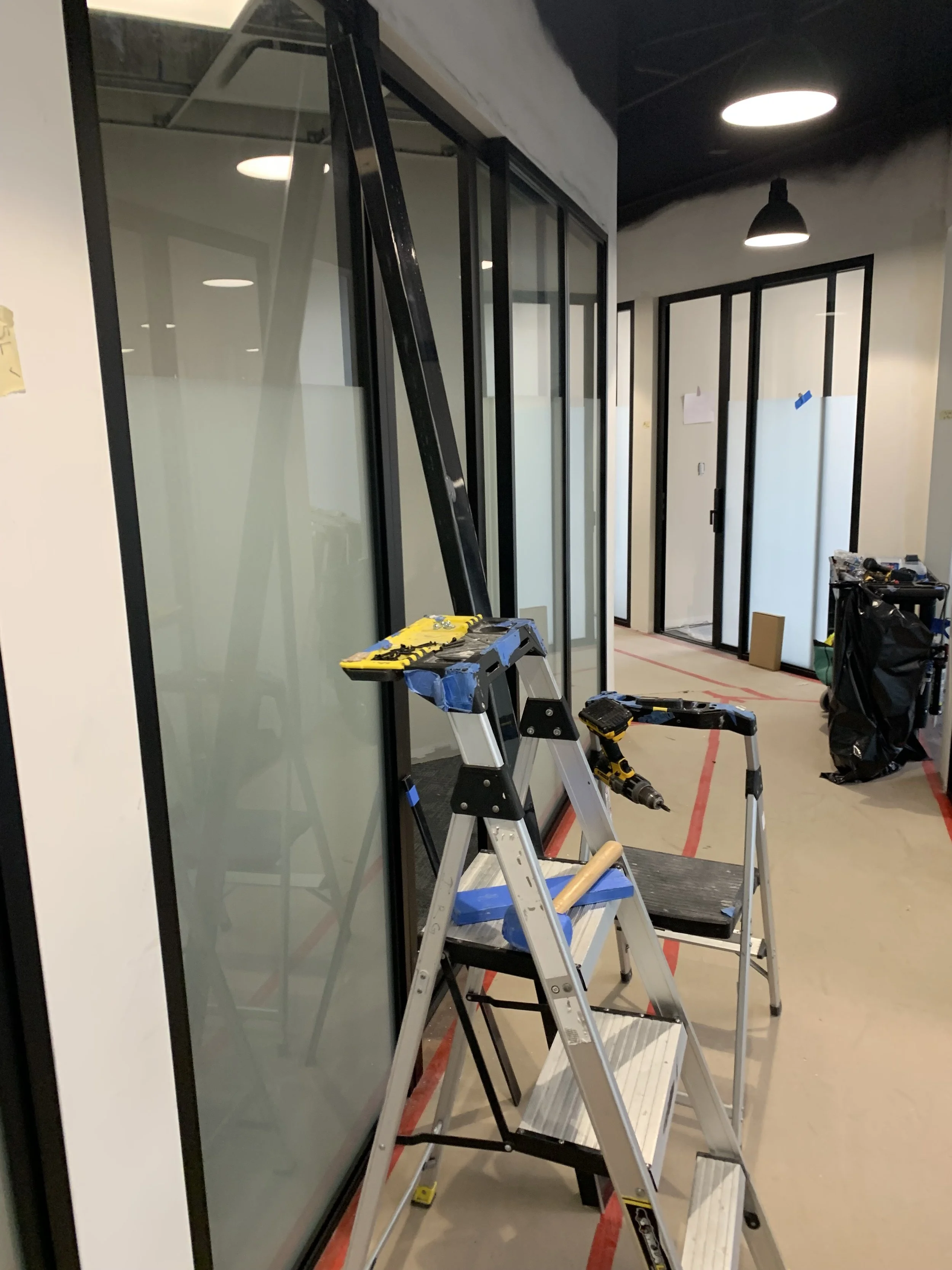 Construction site in an office with a glass partition wall, ladders, tools, and unfinished walls.