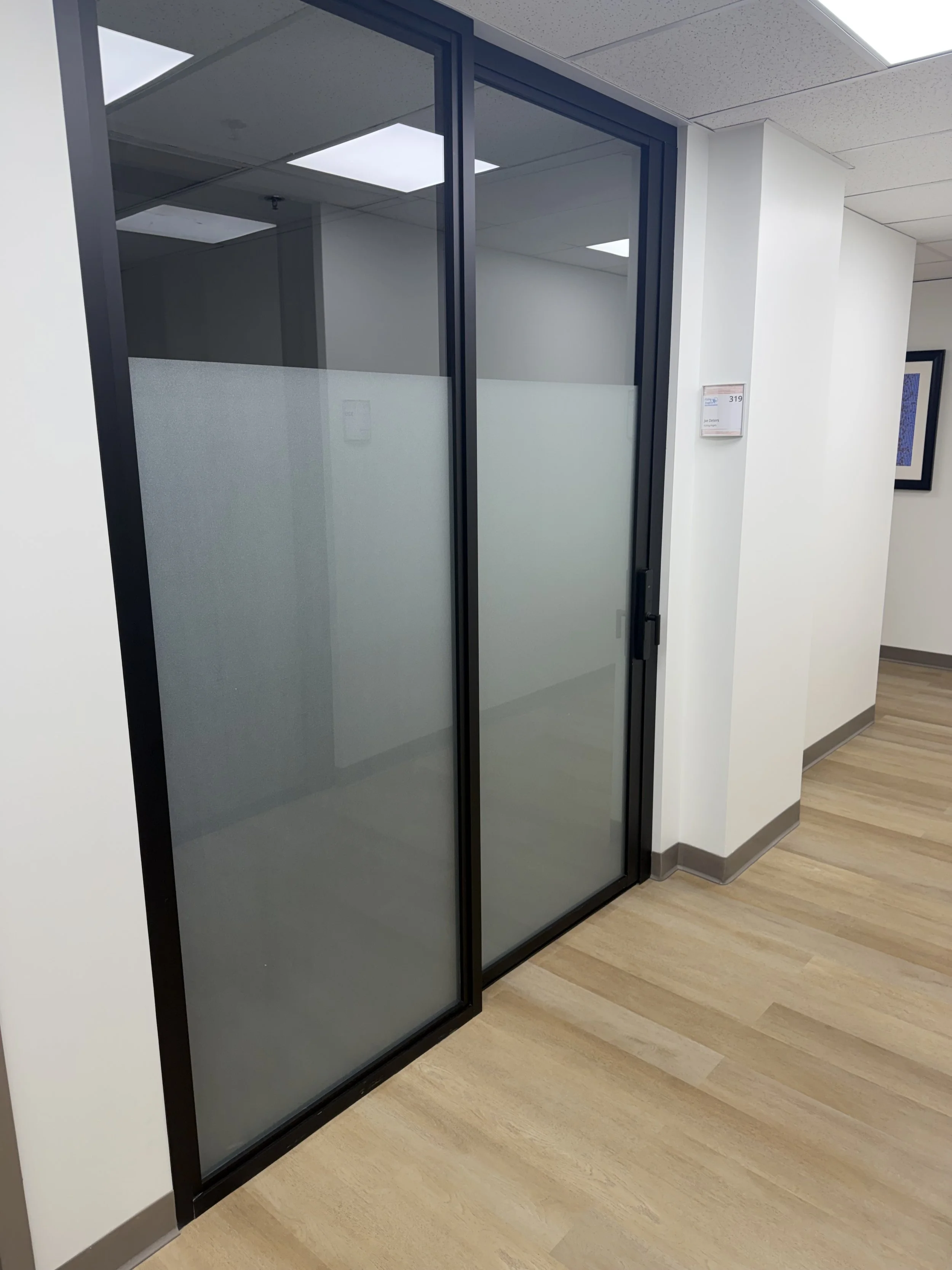 Frosted glass office door with black frame and handle, located in a modern office hallway with white walls, a beige wood floor, and ceiling lights.