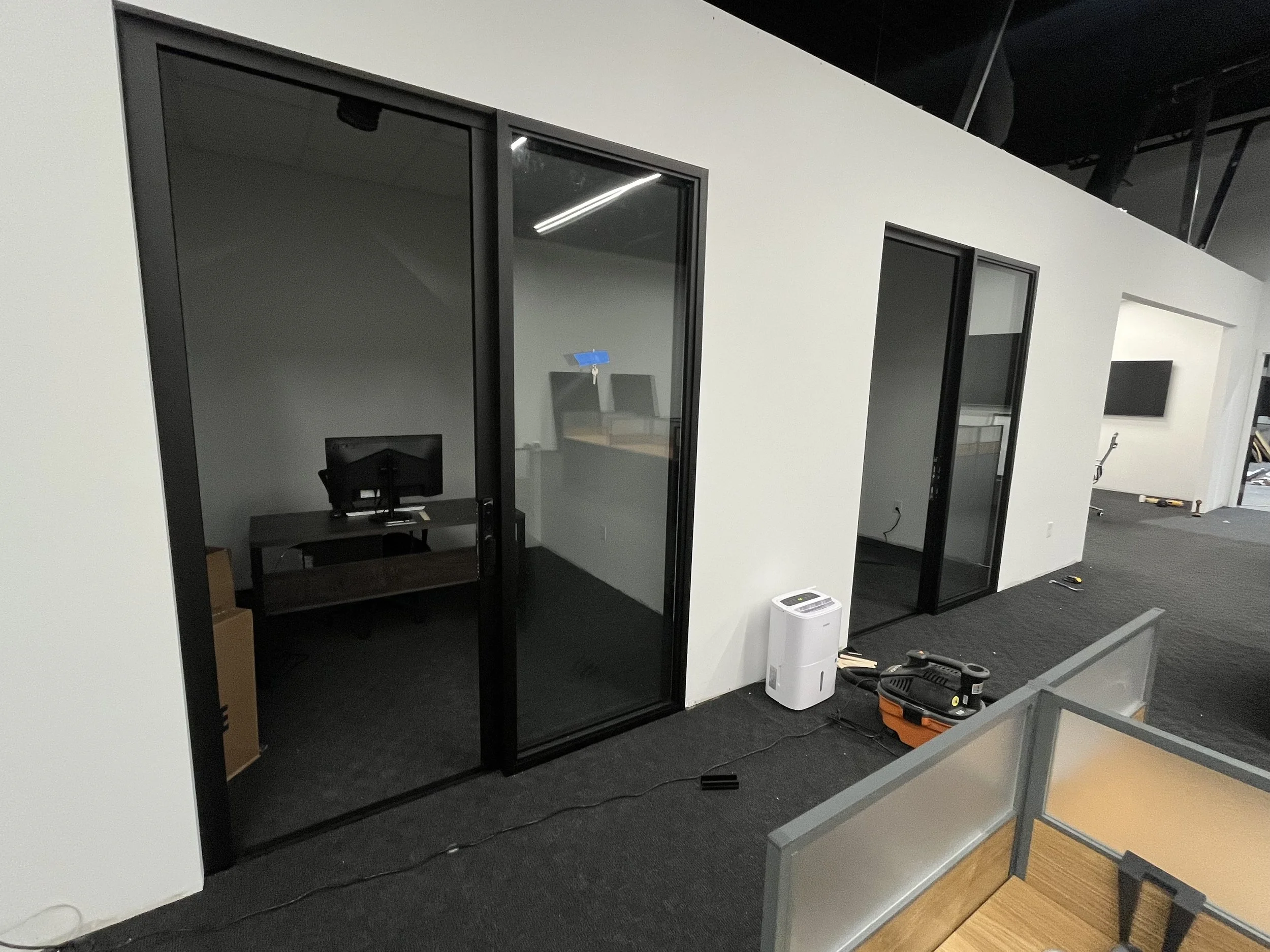 Office space with glass-walled conference rooms, a desk with a computer, a white air purifier, and construction tools on the carpeted floor.