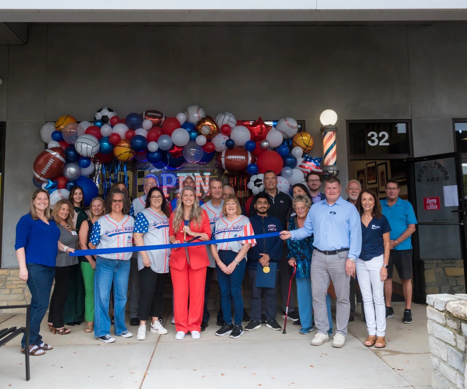 Group of people gathered outside a building decorated with balloons, celebrating an event, with a ribbon-cutting ceremony led by a woman in a bright red suit.