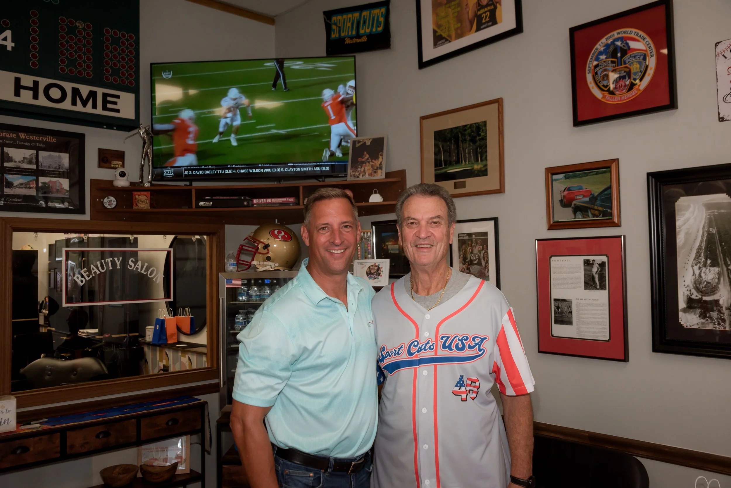 Two men smiling and standing together in front of a wall decorated with sports and vintage memorabilia. One man is wearing a light blue shirt, and the other is wearing a white jersey with red, white, and blue details.