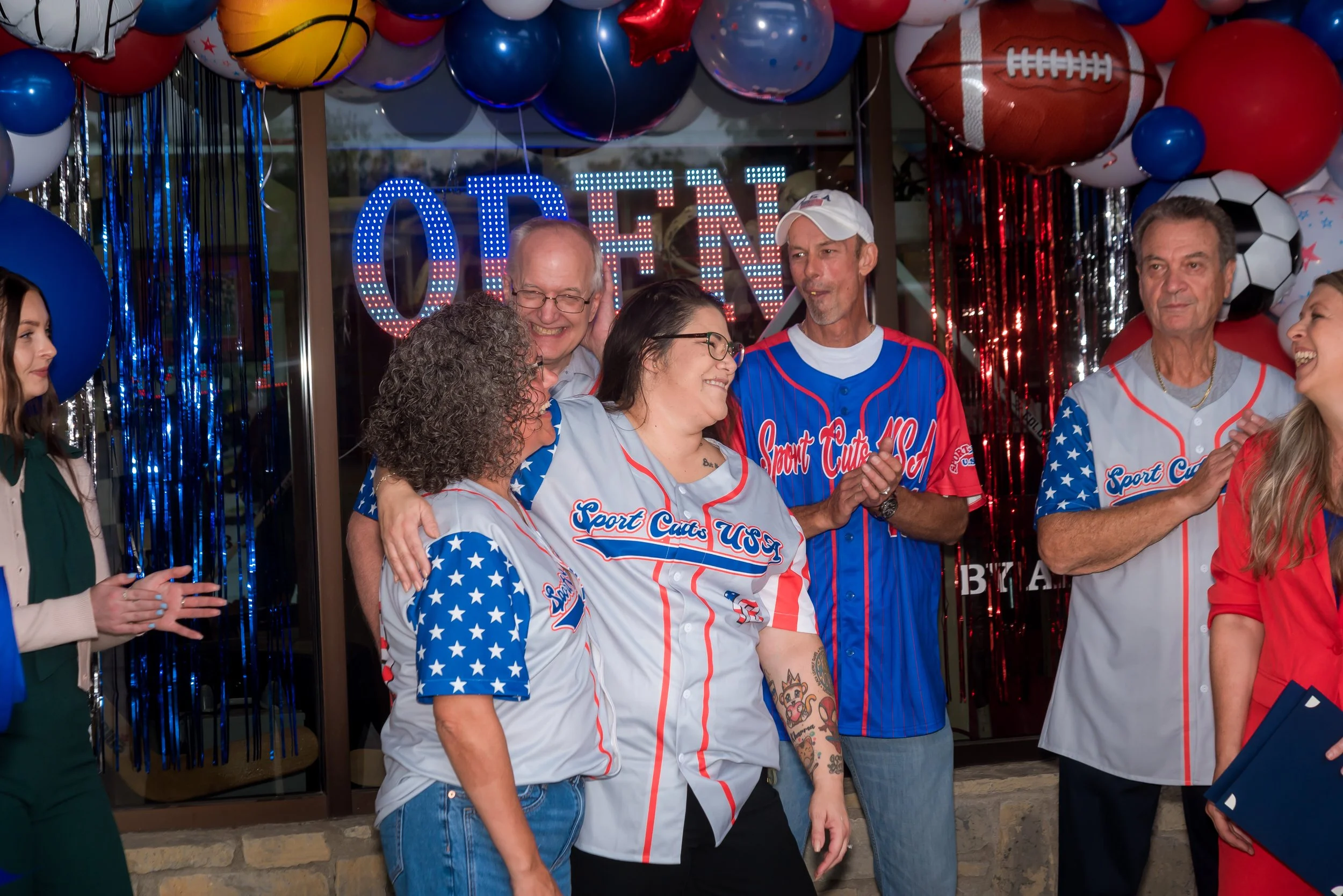 A group of people celebrating at a party decorated with patriotic and sports-themed balloons, including baseballs, footballs, and soccer balls, with a large illuminated 'OPEN' sign in the background.