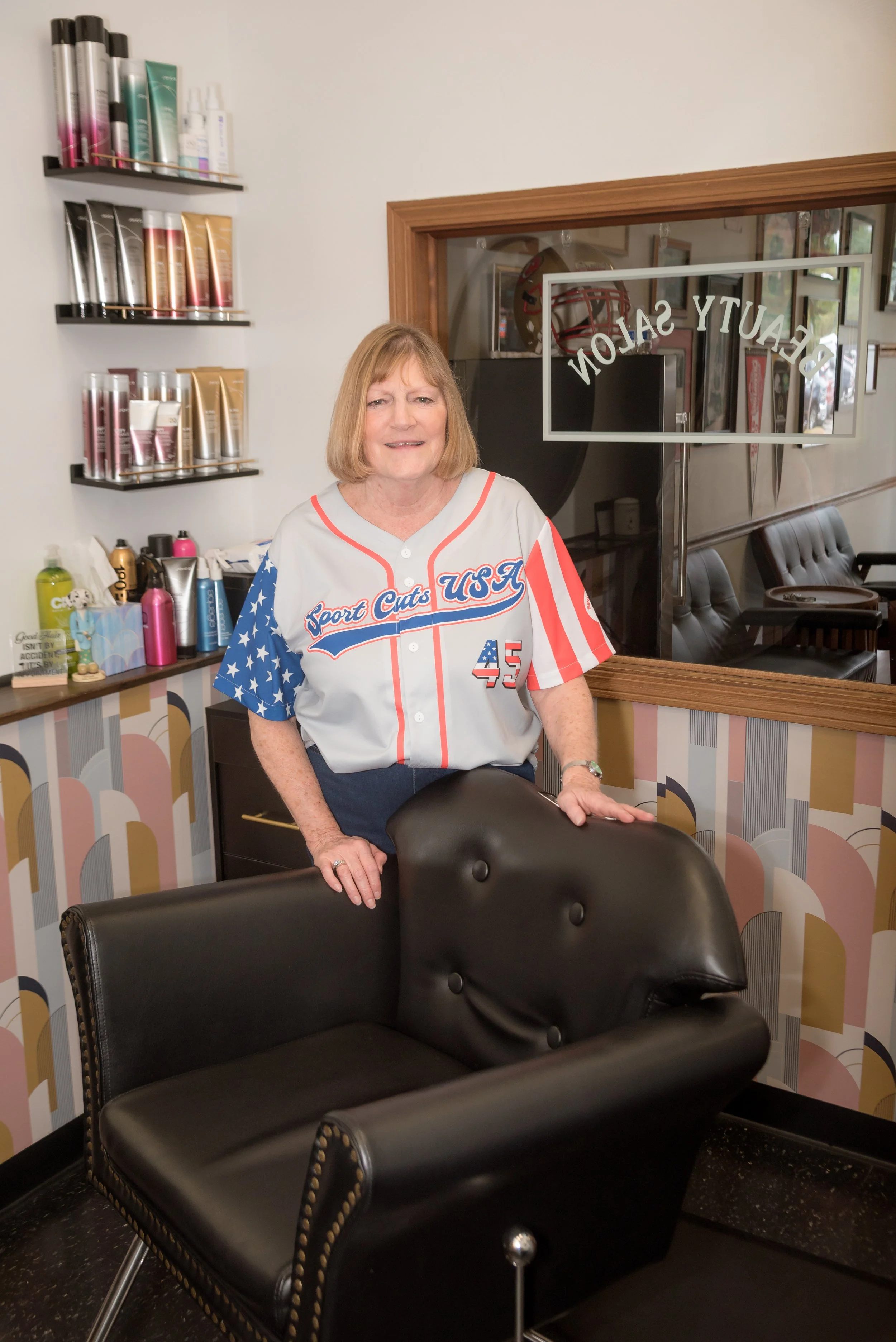 A woman in a USA sports jersey standing in a salon, with black salon chairs in front of her and shelves of hair products on the wall to her left, behind a wooden framed glass window with the salon name.