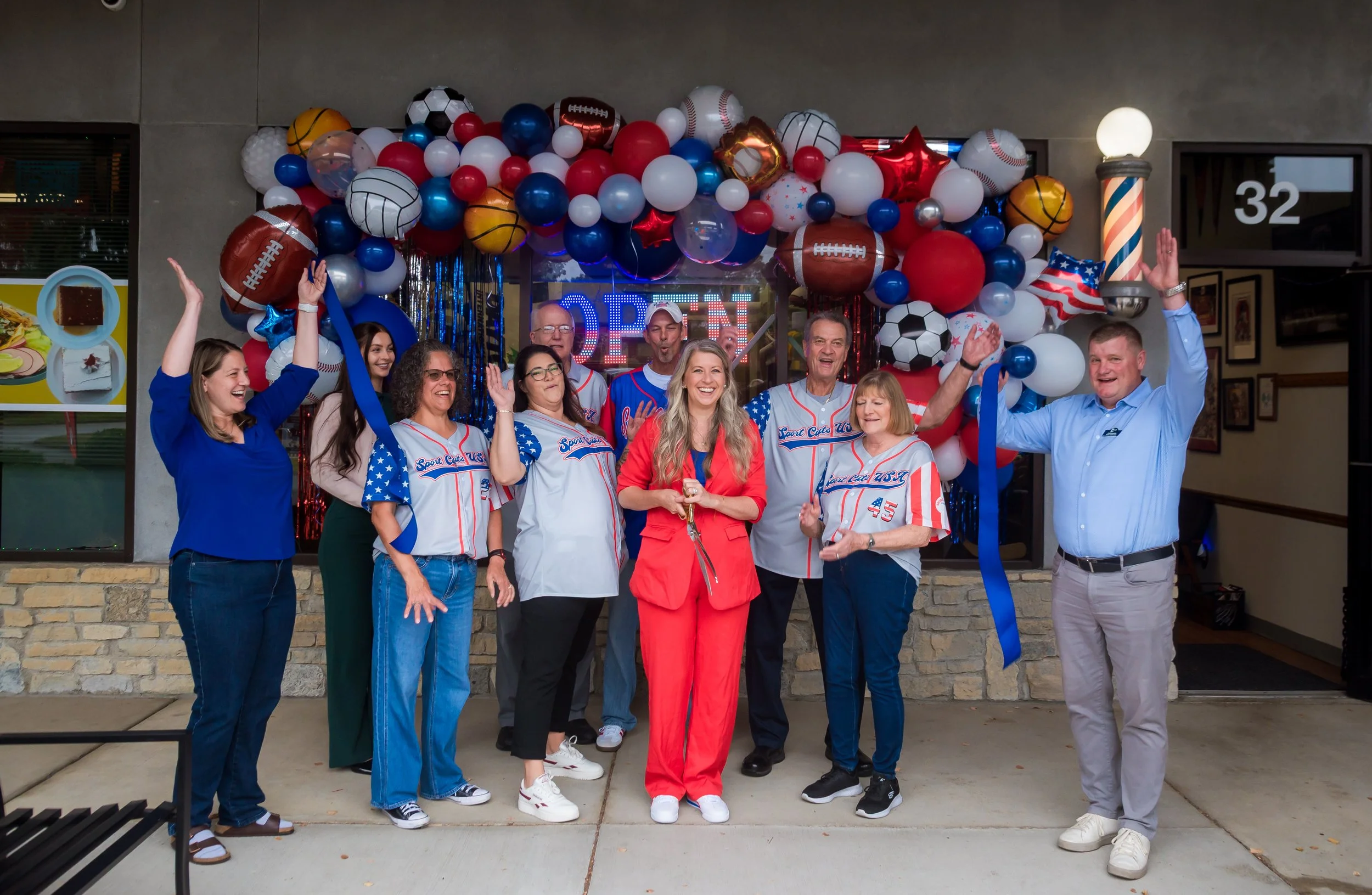Group of people celebrating New Ownership and 35 years of serving the Westerville OH front of Sport Cuts USA storefront with balloons, wearing sports jerseys and casual clothes, smiling and raising their hands.