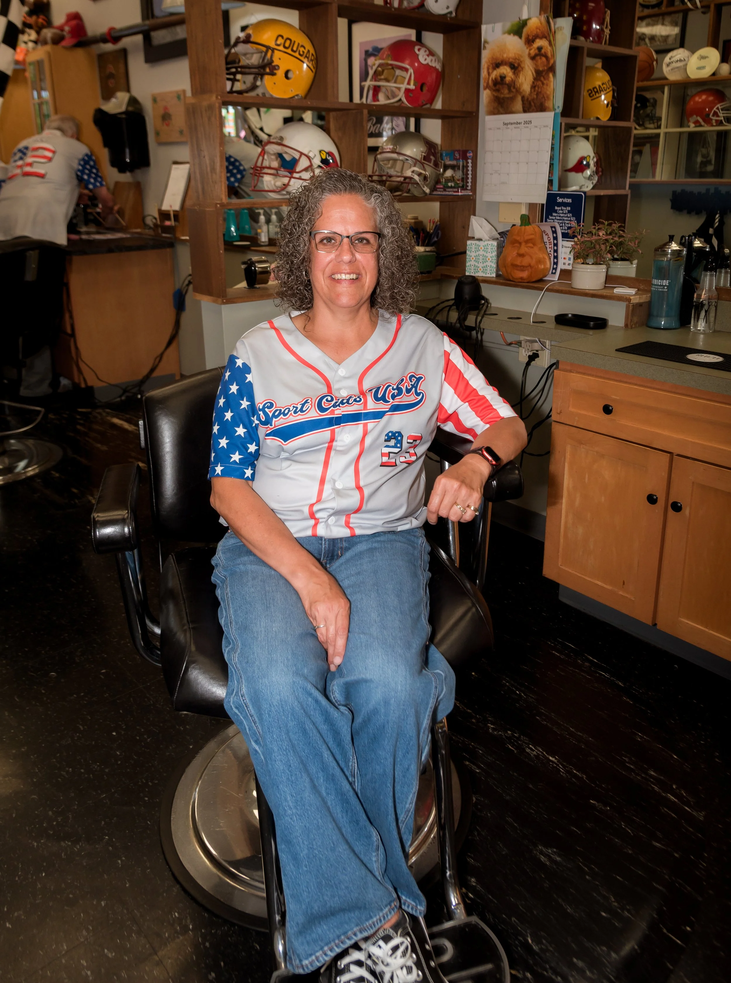 Christie a Barber wearing a jersey with stars and stripes, sitting in a barber chair inside a sports-themed barbershop, smiling at the camera.