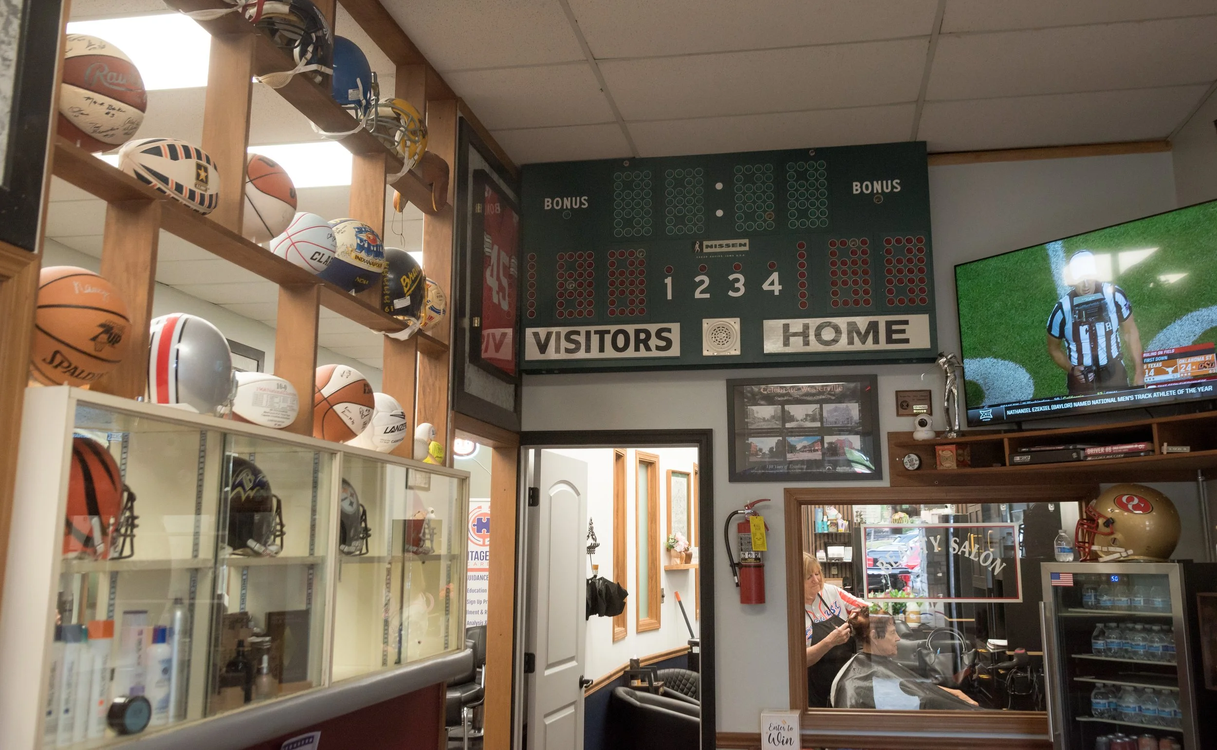 Indoor sports memorabilia store with wooden shelves displaying various footballs, a scoreboard on the wall, and people sitting inside, with a woman seated near the window.