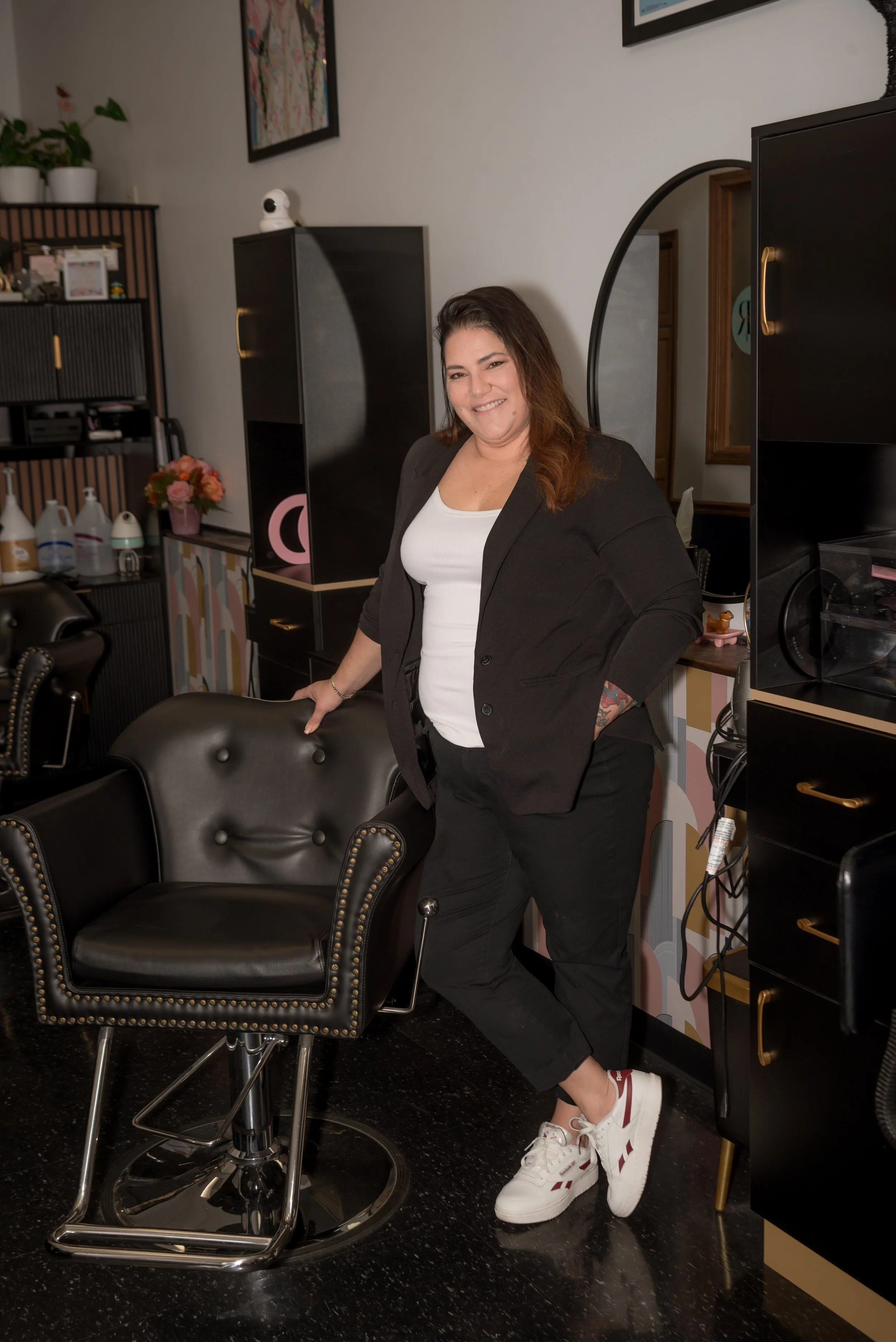 A woman with brown hair, black blazer, white shirt, and black pants standing beside black salon chairs and furniture in a salon or beauty parlor, smiling at the camera.