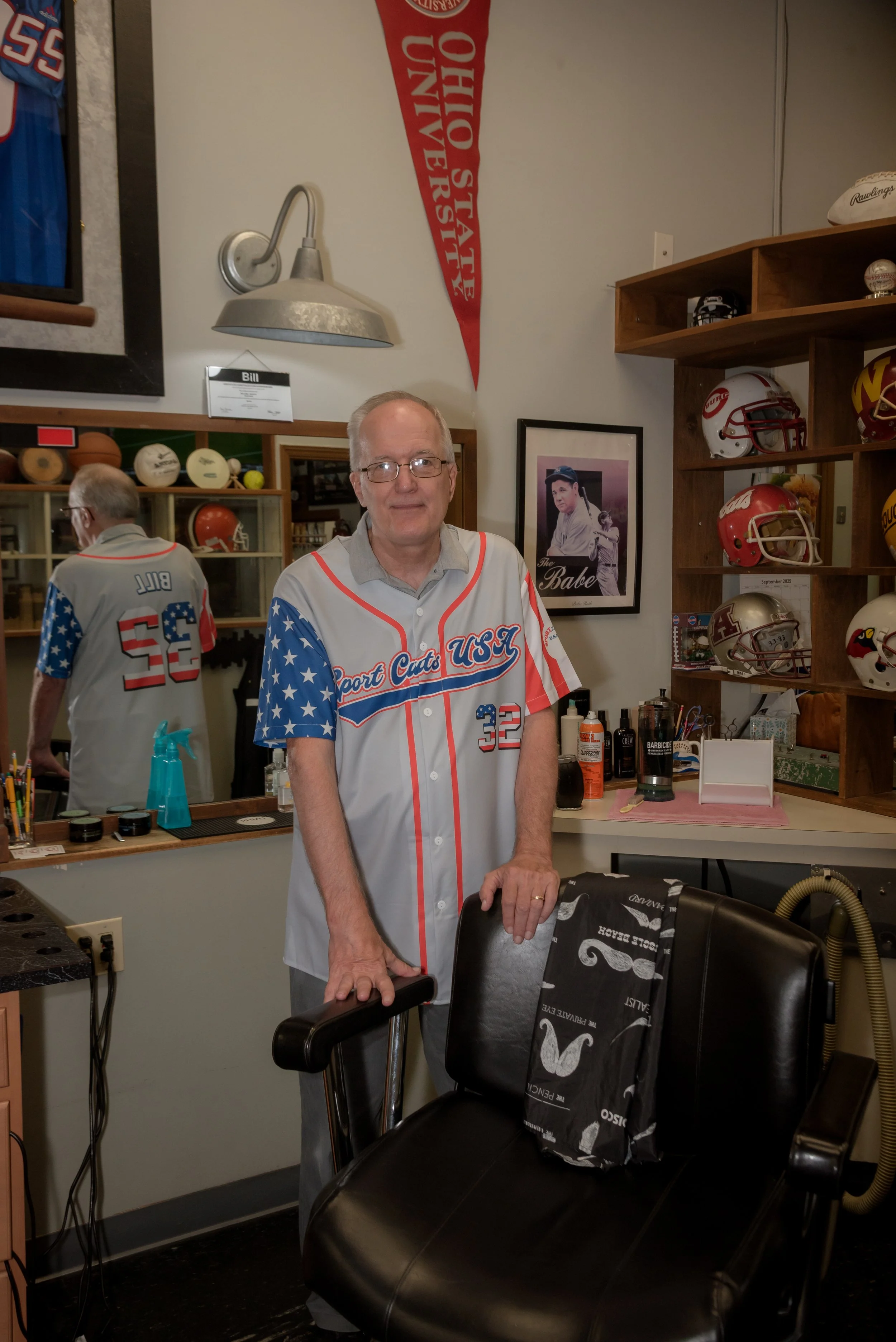 A man wearing a USA sports team jersey standing in a barbershop, with barber tools on the counter, framed photos and sports memorabilia on the wall, and a black barber chair in front of him.