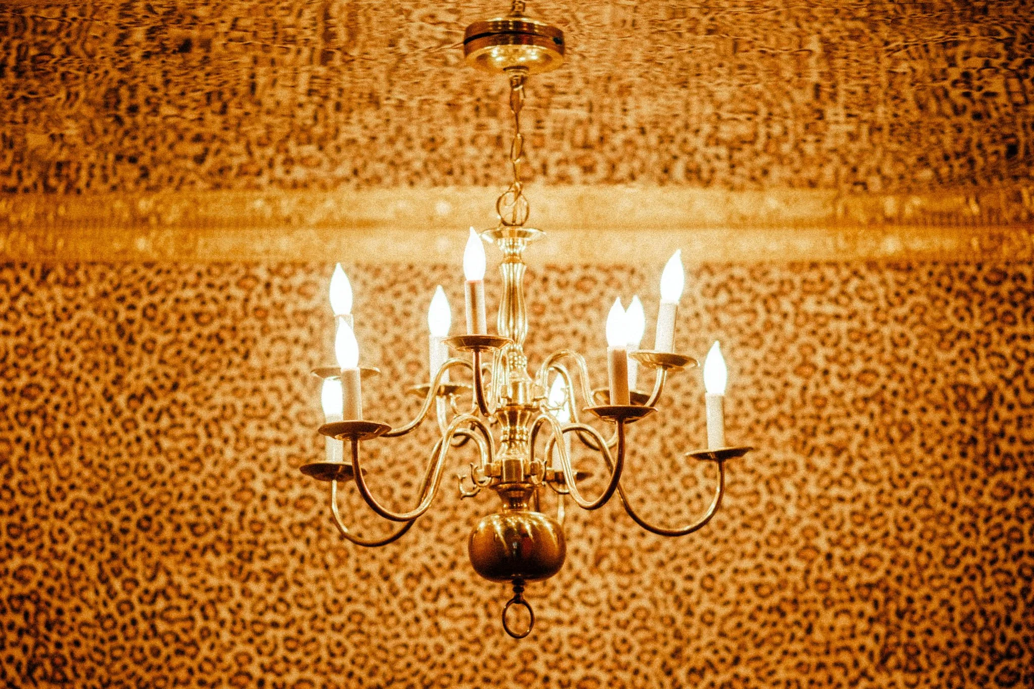 A brass chandelier with lit candles hanging from a gold chain, set against a textured, leopard-print wallpaper.