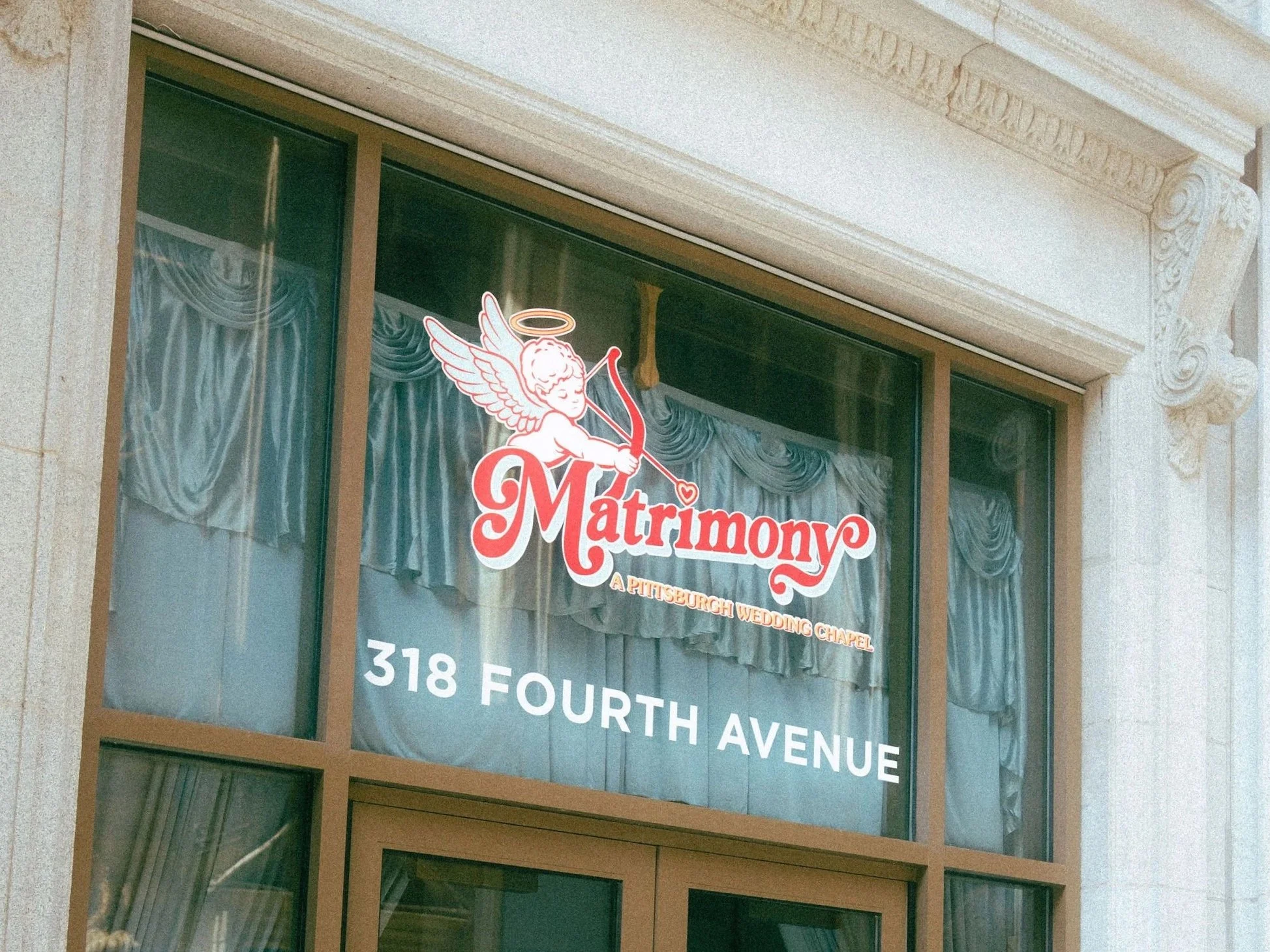 Sign for a marriage chapel featuring a cartoon angel with a halo and bow and arrow, with the words "Marriage" and "A Pittsburgh Wedding Chapel" on the window.