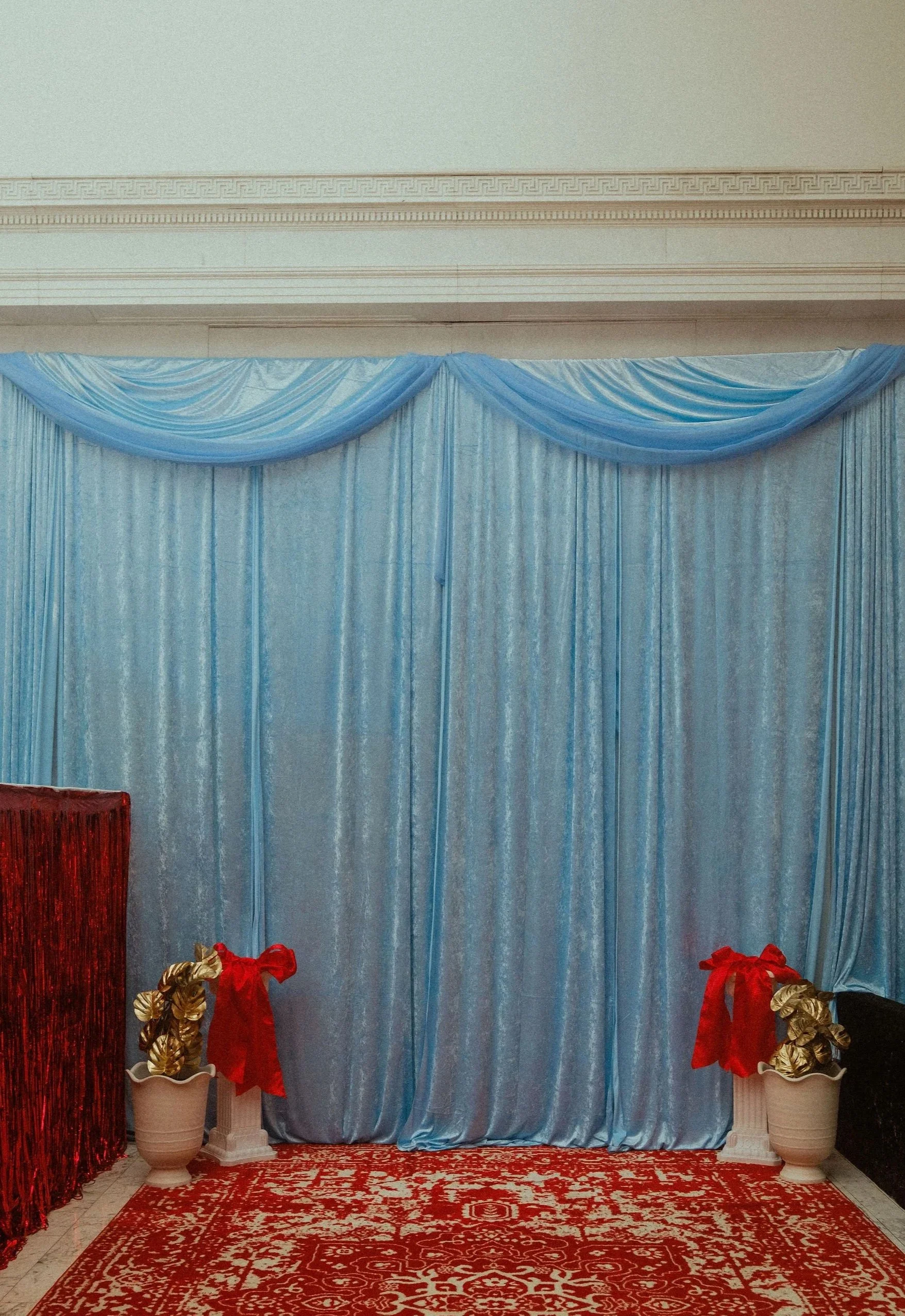 Decorative backdrop with light blue curtains and draped fabric at the top, set on a red patterned carpet. Two white flower pots with gold and red gift bags are in front, on either side.